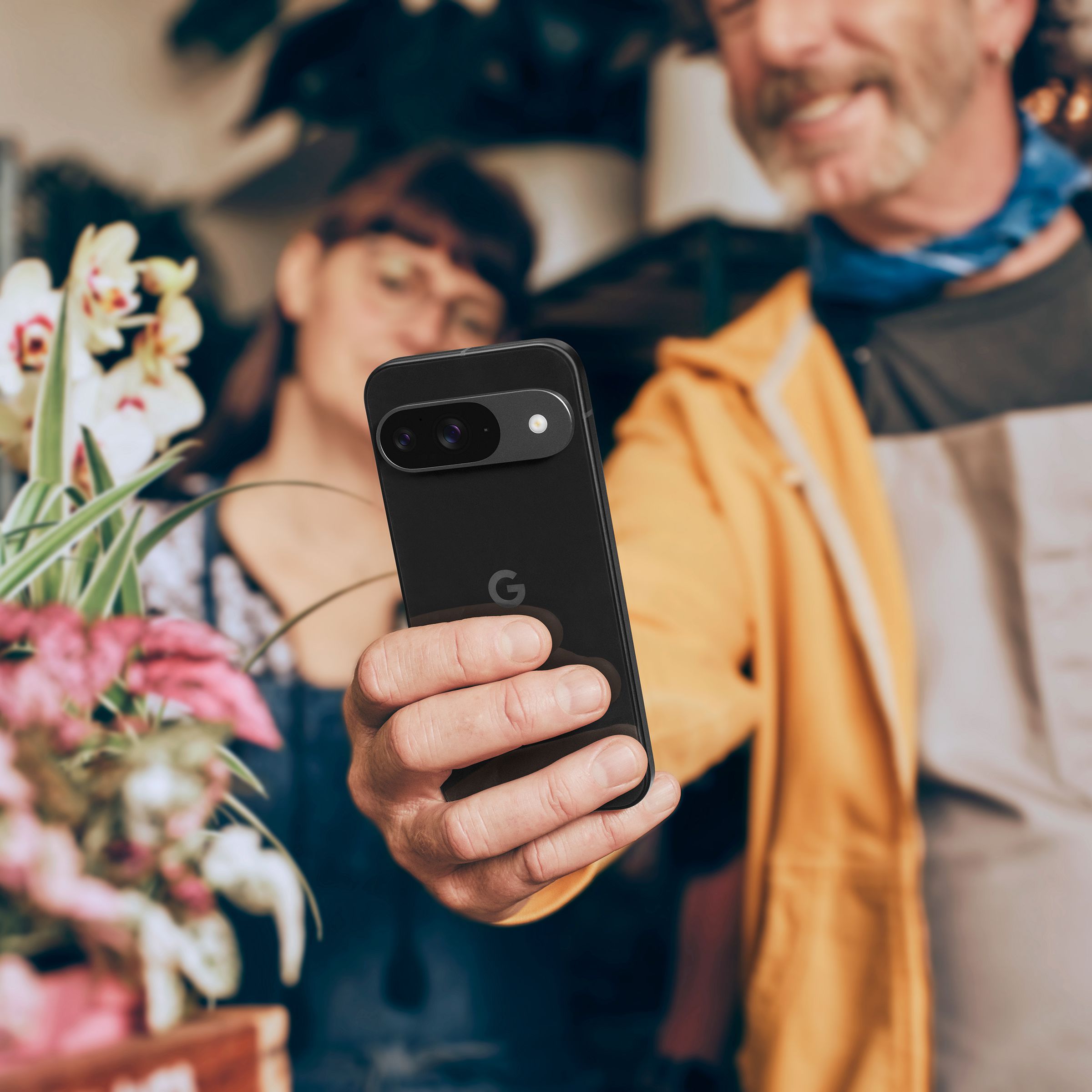 A person holding a Pixel 9 phone up at arm’s length to take a picture of flowers.