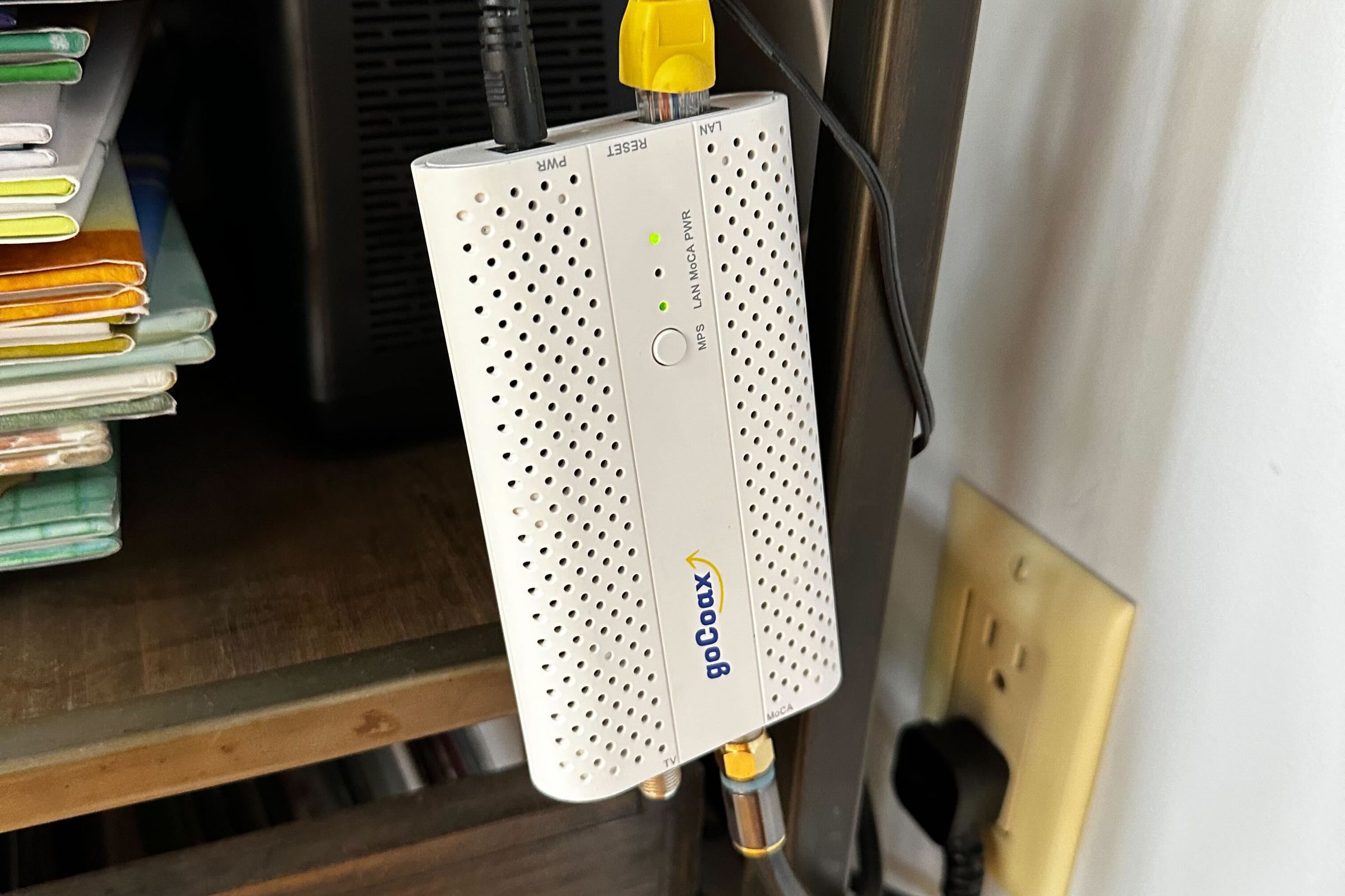 shot of a white MoCA adapter labelled “goCoax” with ethernet and power cables emerging from the top and a coax cable coming from the bottom. The adapter is dangling in midair in front of a bookshelf, with a power outlet visible in the background. 
