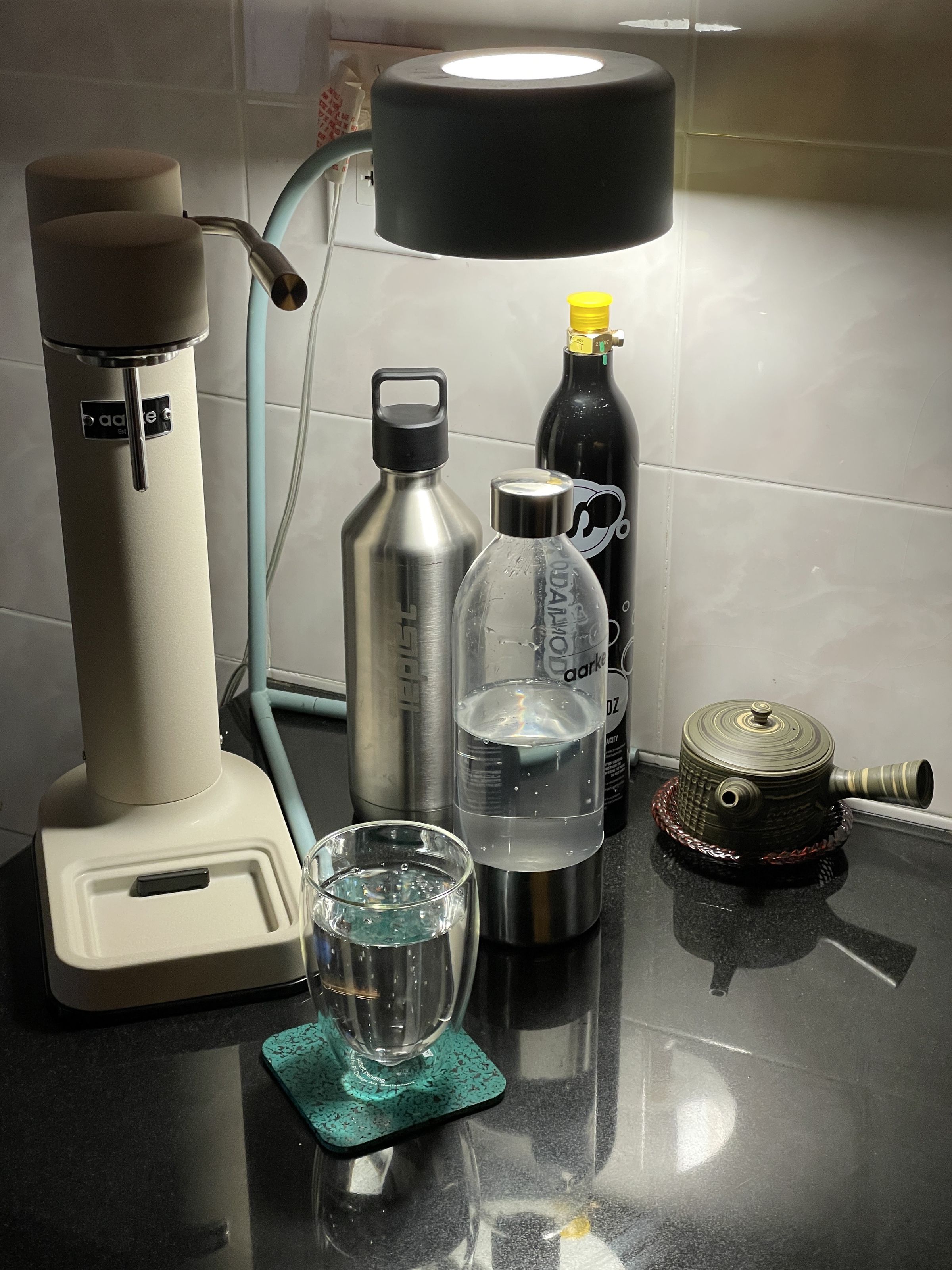 An image of an Aarke Carbonator III, a bottle full of carbonated water, a steel travel bottle for seltzer, a double walled Pavina glass by bodiun and a nerikomi-style clay teapot that is unrelated to everything else.