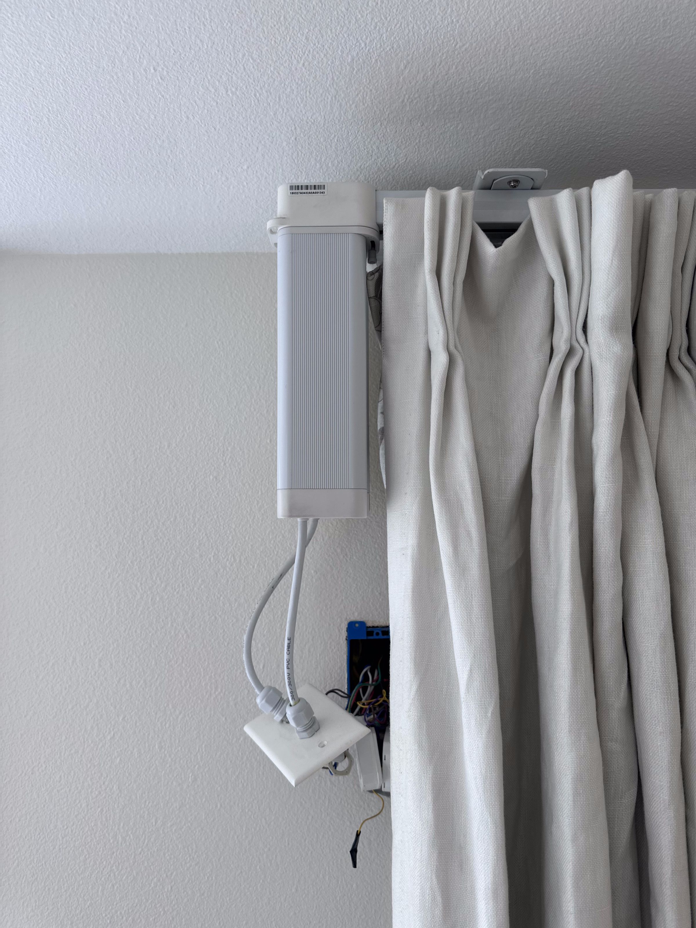 An image of a large white box with two cables coming out of it. It is attached to a curtain and appears to be a motor to open and close the curtains.