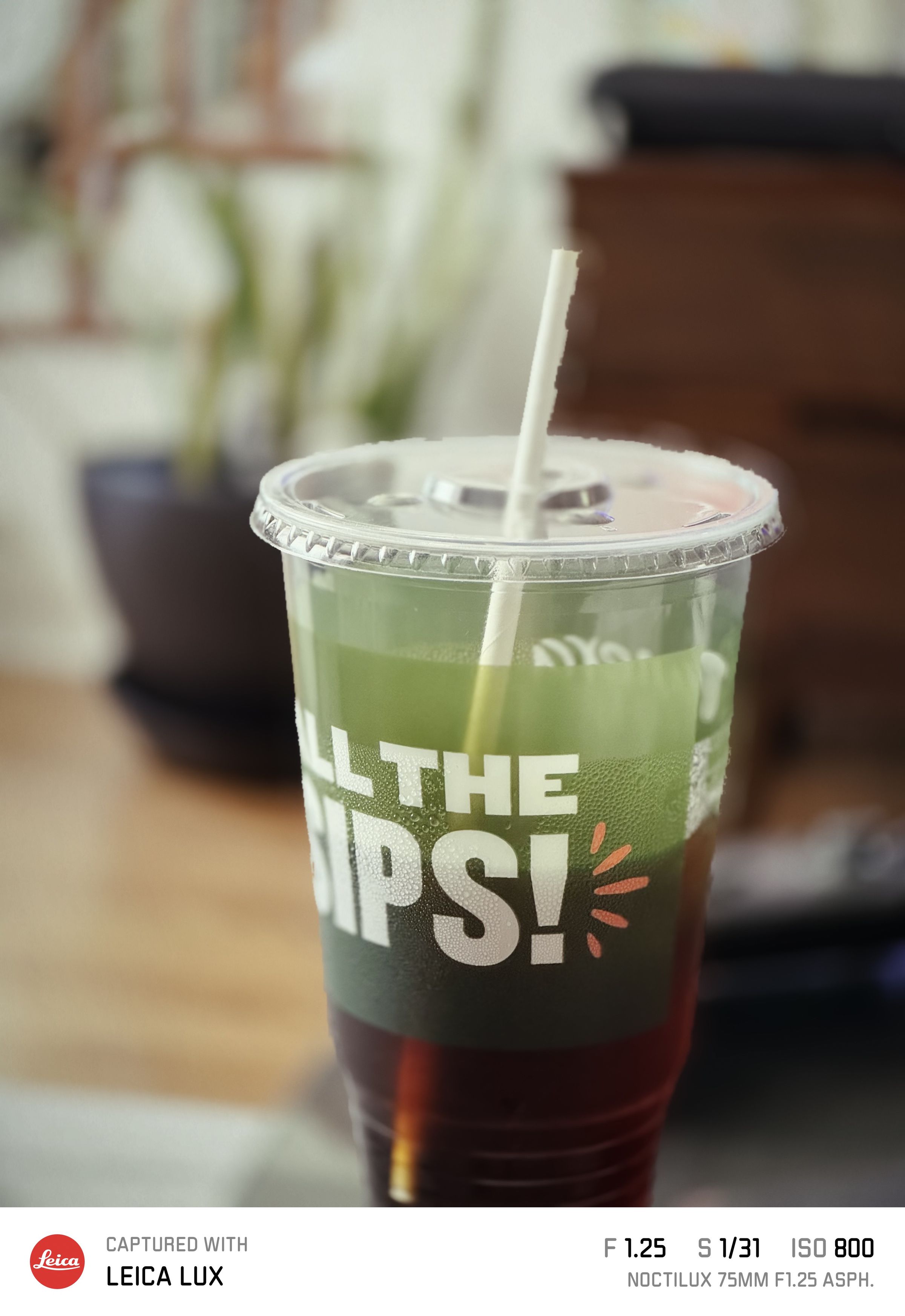A portrait mode photo taken of an iced coffee cup with a straw. The photo also has a branded Leica Lux frame showing its metadata.
