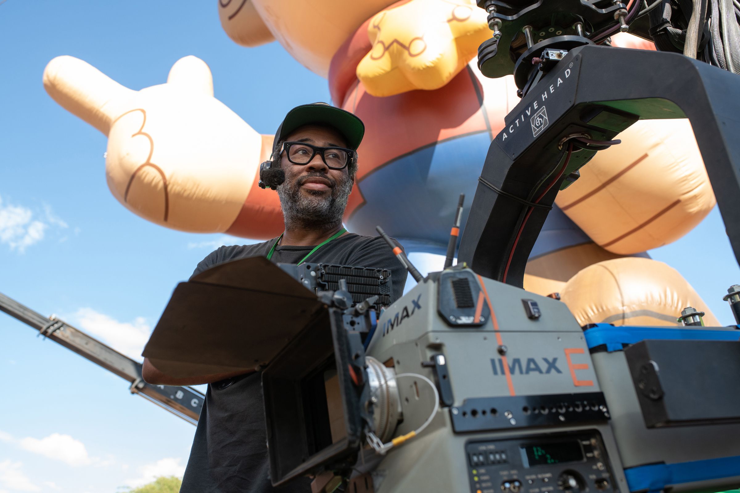 Jordan Peele on the set of Nope standing behind an IMAX camera, and in front of a massive, inflatable cartoon cowboy.