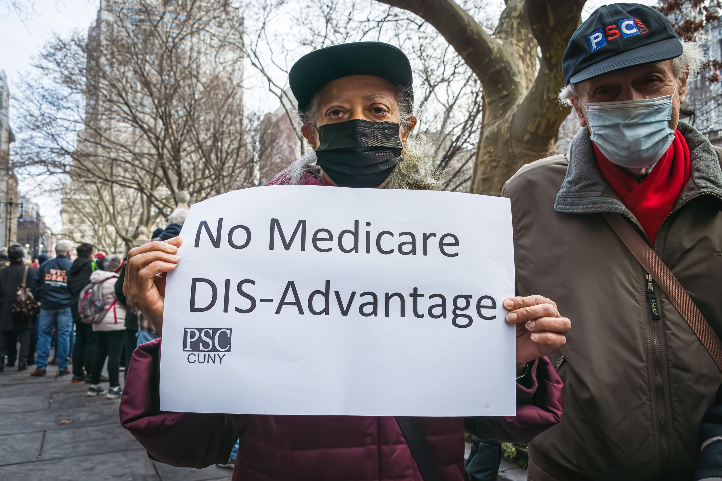 Demonstrator with mask holding sign reading “No Medicare Dis-Advantage.”
