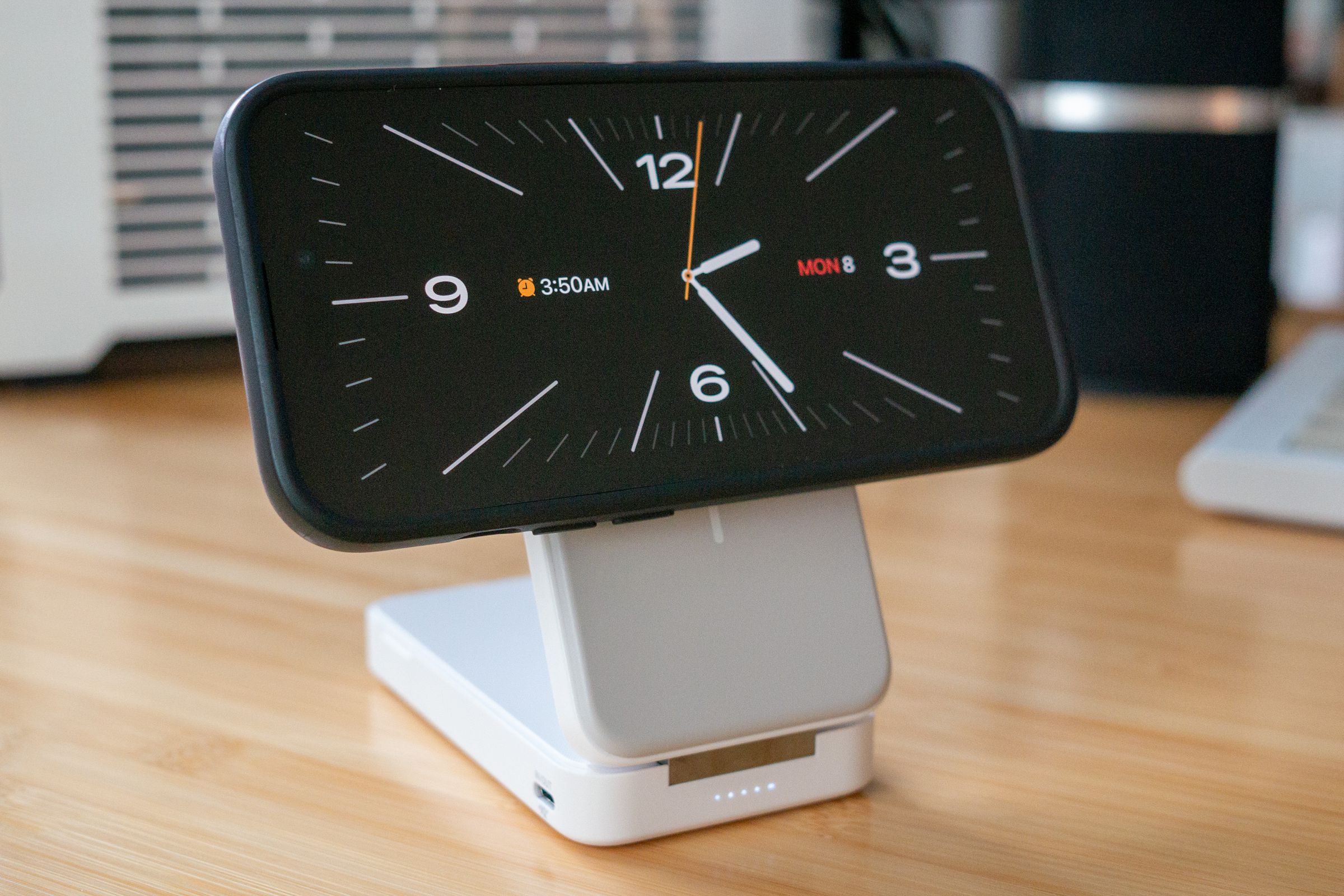 A white-and-grey folding slab charger holding an iPhone 14 Pro on its Qi2 magnetic charging surface. The iPhone is in StandBy mode and displaying a white-on-black analog clock