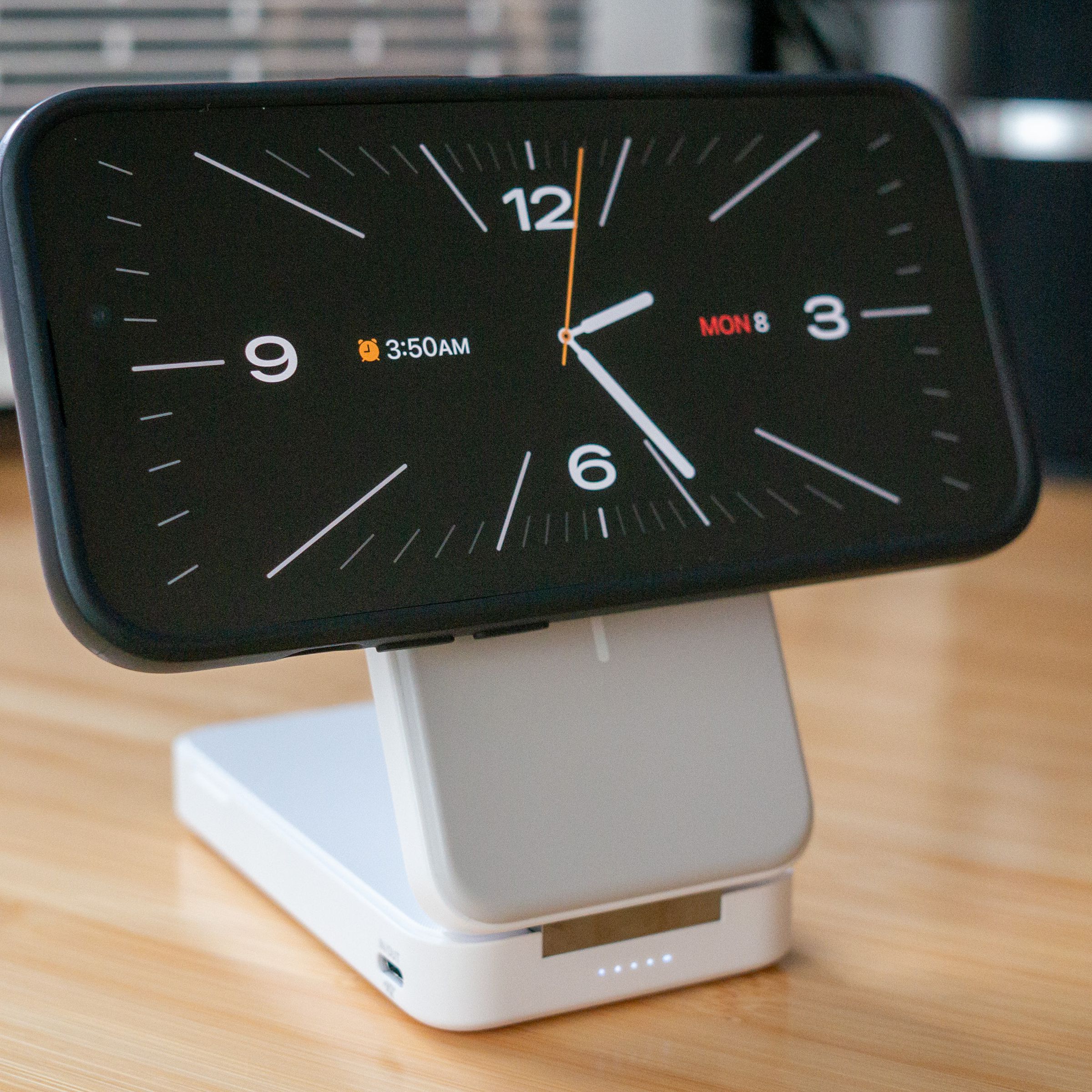 A white-and-grey folding slab charger holding an iPhone 14 Pro on its Qi2 magnetic charging surface. The iPhone is in StandBy mode and displaying a white-on-black analog clock