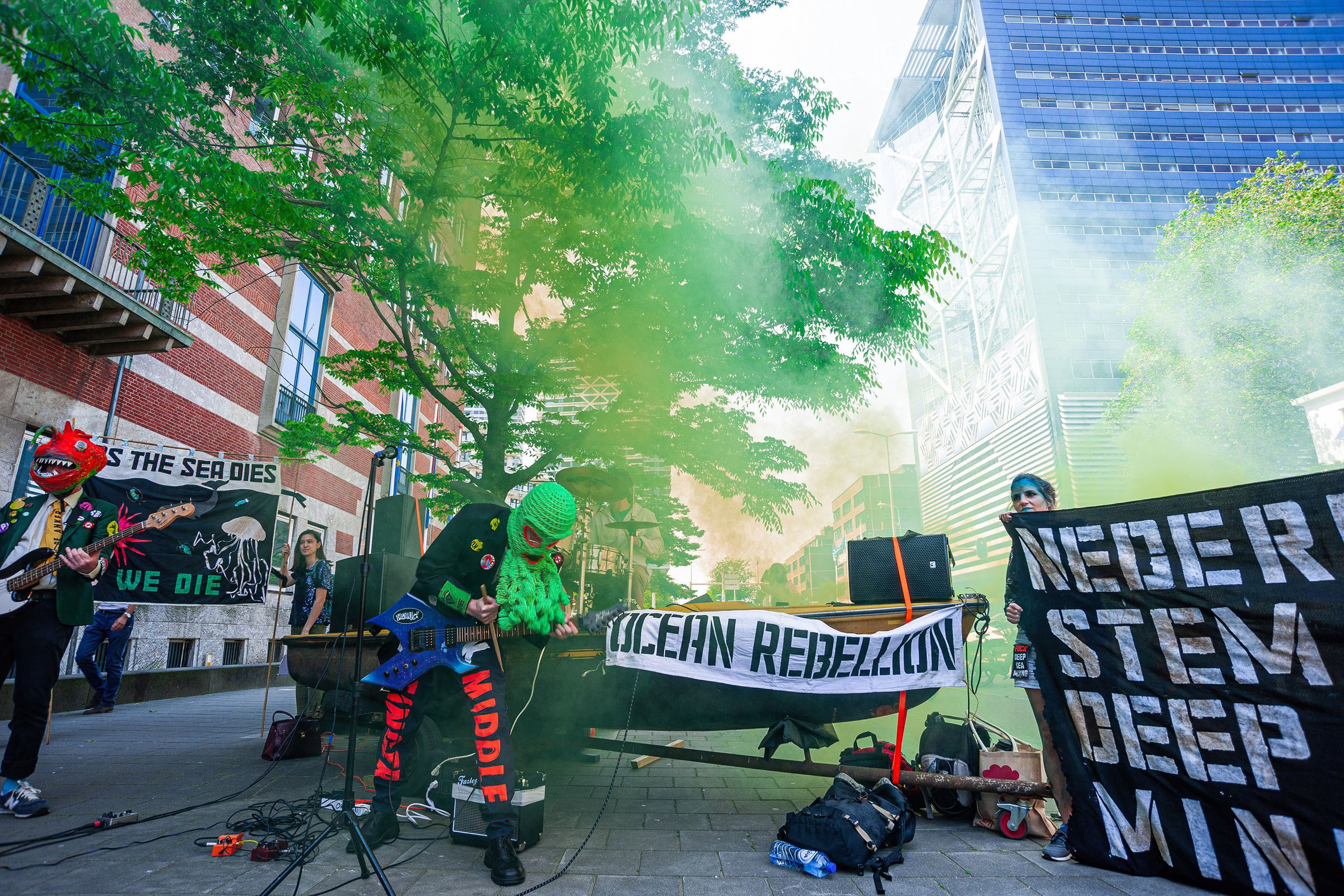 Band members wearing masks play instruments in a haze of green smoke with a banner behiind them that says “ocean rebellion.”