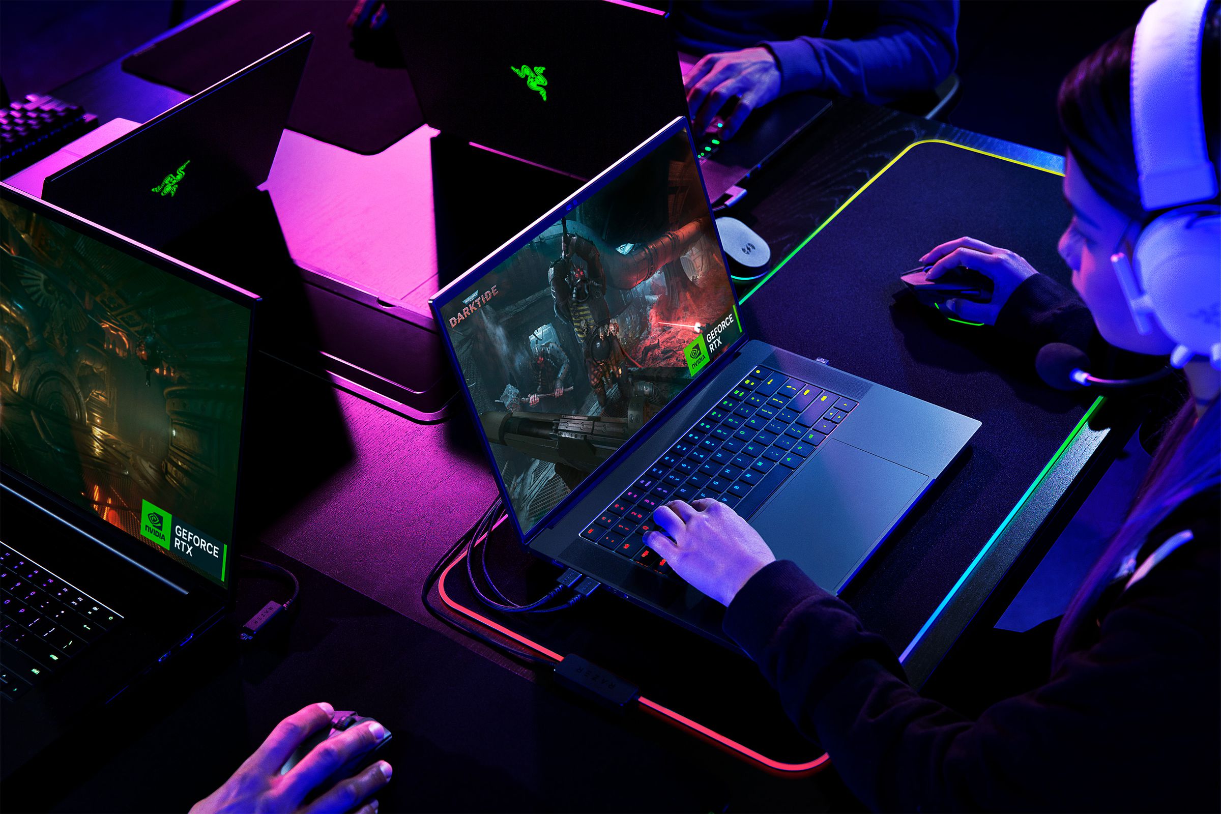 Stylized image with pink and blue lighting of several gamers at a table. All are using Razer Blade laptops with large desk mats plugged into them. The desk mats have RGB edge lighting and are wirelessly powering the gaming mice.