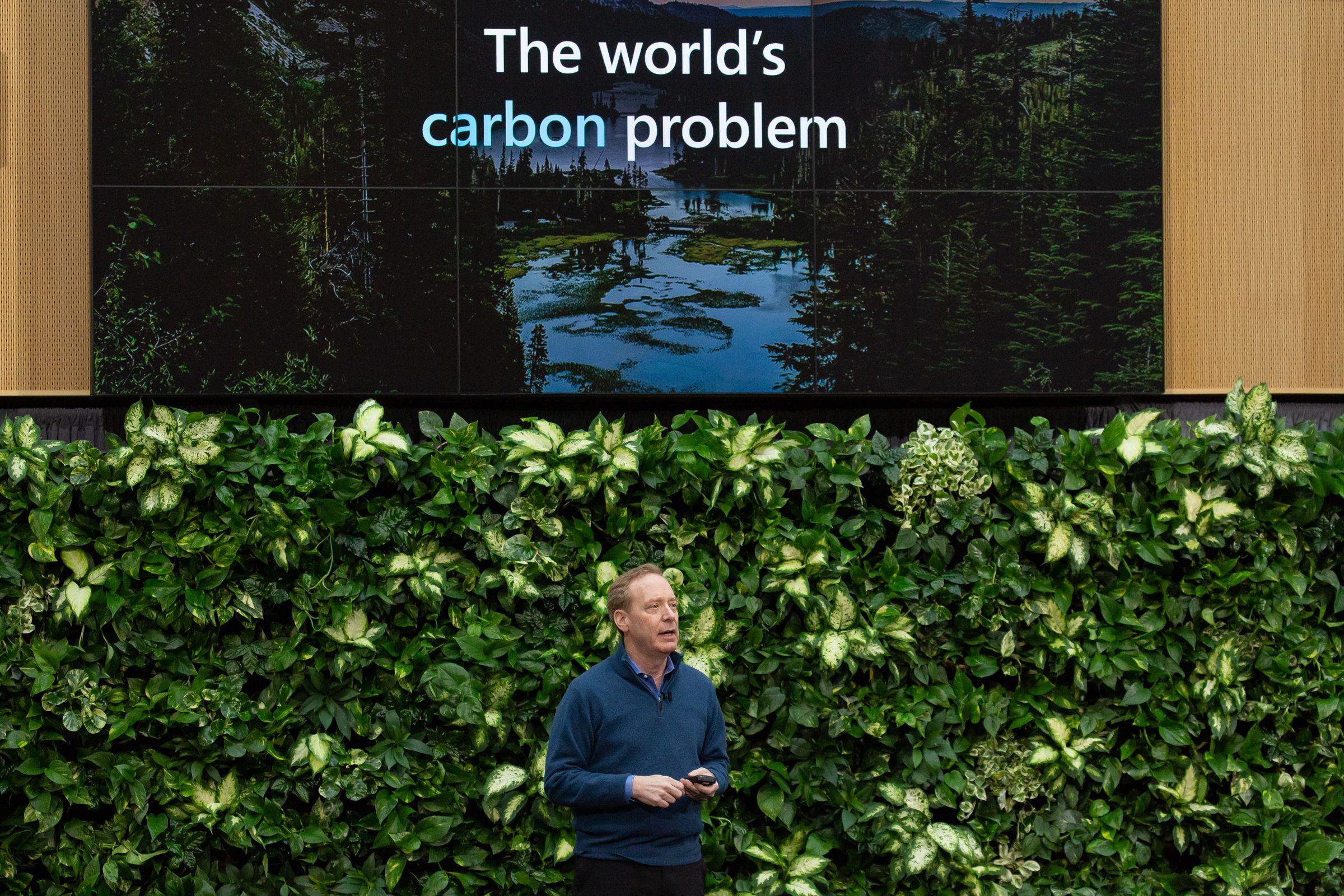 A person stands in front of a wall of greenery. Above him, a screen says “the world’s carbon problem.”