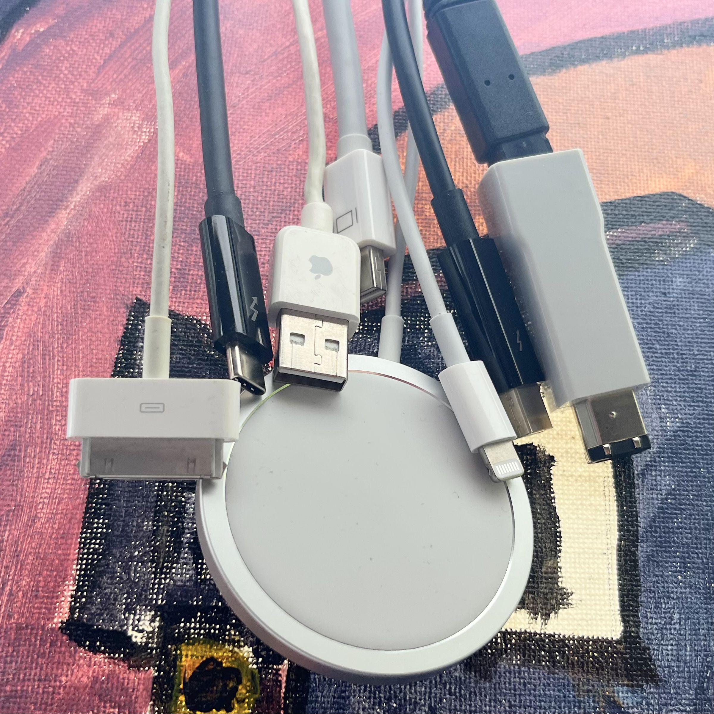30-pin, usb-c thunderbolt 3, usb a with apple logo, magsafe for iphone, mini DisplayPort, lightning, thunderbolt 2, firewire 400 and 800 cables all dangling in front of a purple hue painting