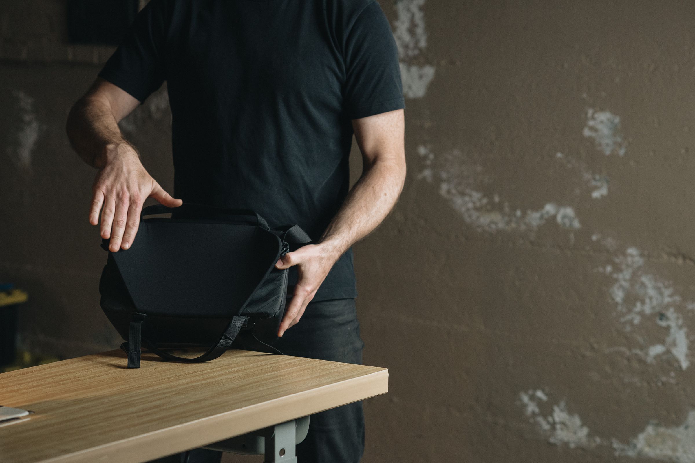 A man demonstrating the flexible, winged back panel of the Moment Rugged Camera Sling bag on a wooden table.