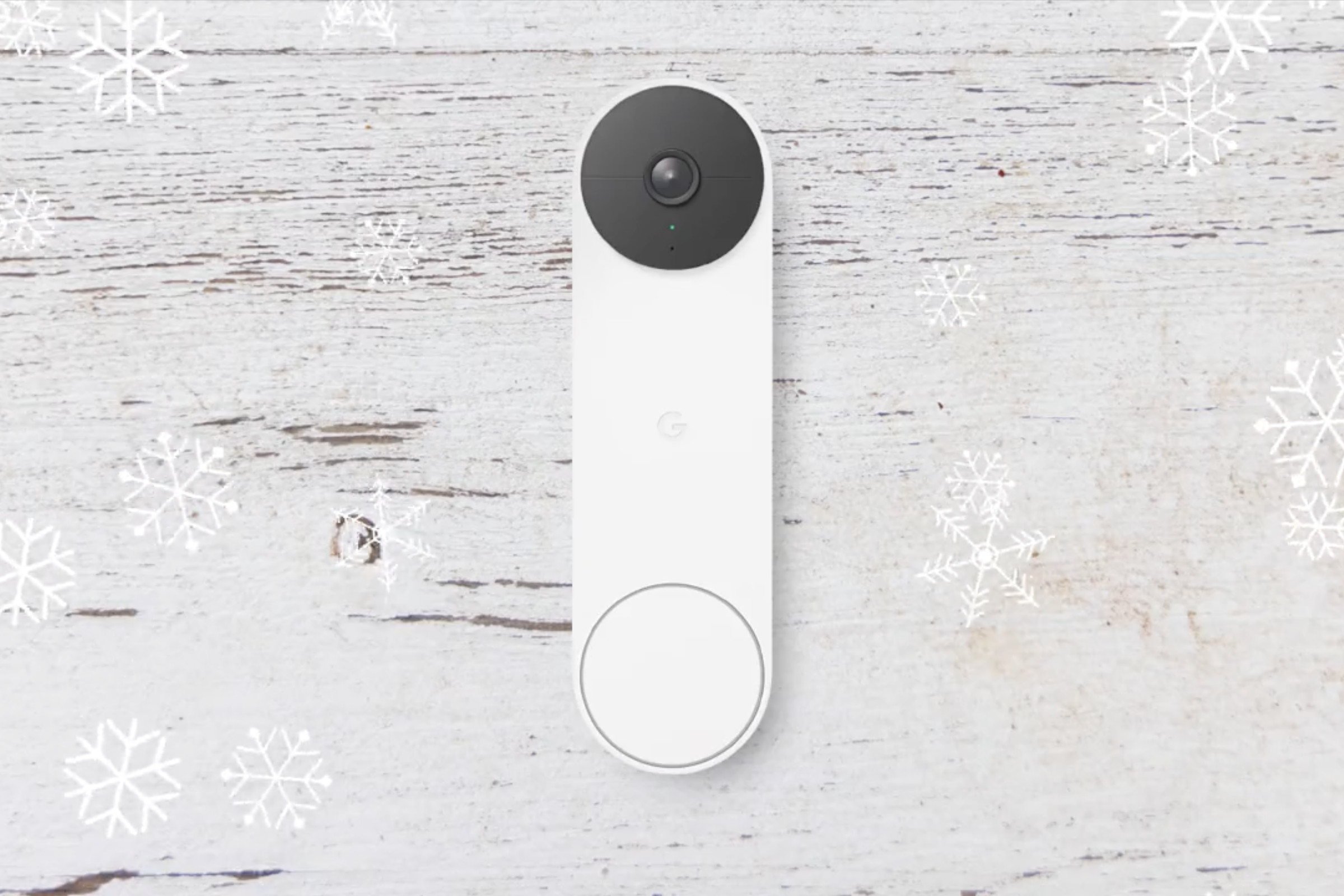 Google Nest doorbell on a gray wood plank. It has a camera lens on the top and a circle button on the bottom. Snowflake designs around the background