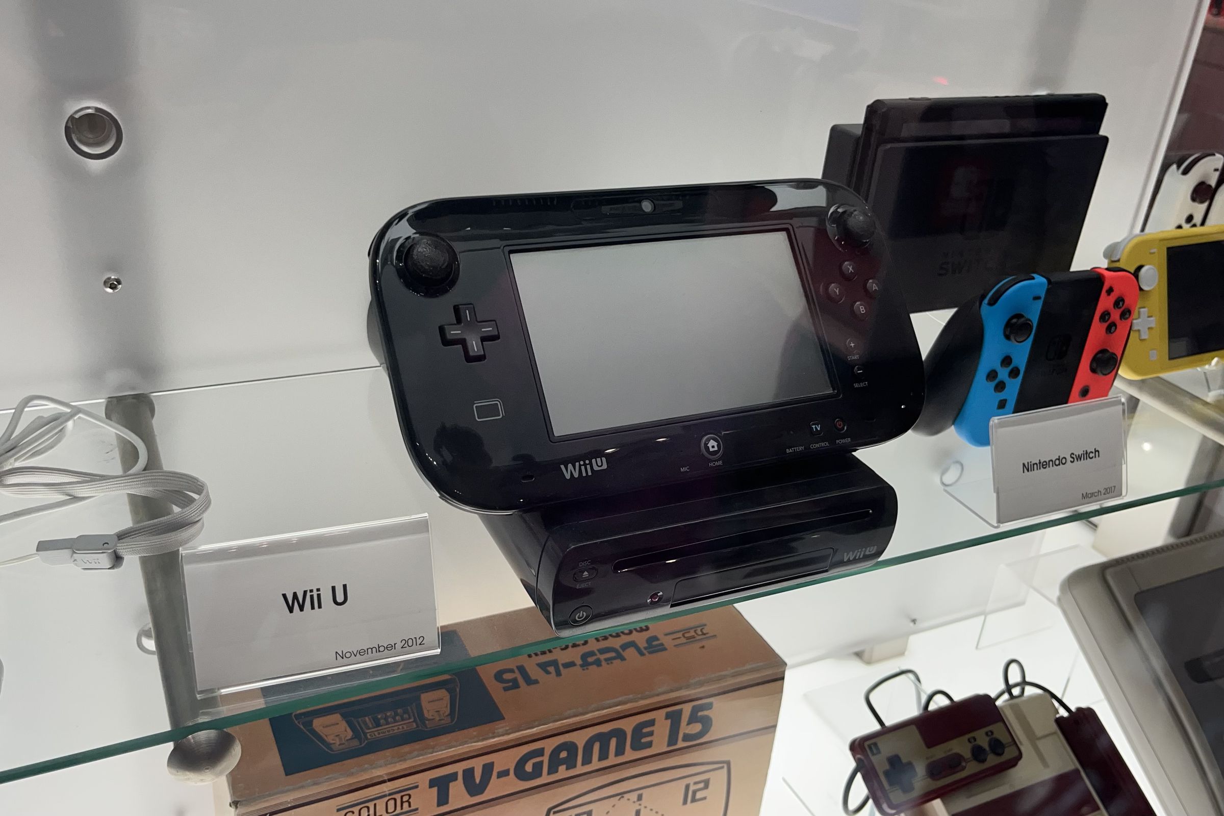 black Wii U system in a glass display case with the Nintendo Switch beside it and the tv-game 15 under it. The Wii U gamepad with screen is sitting on top of the Wii U system. a famicom and super famicom are on the bottom right.
