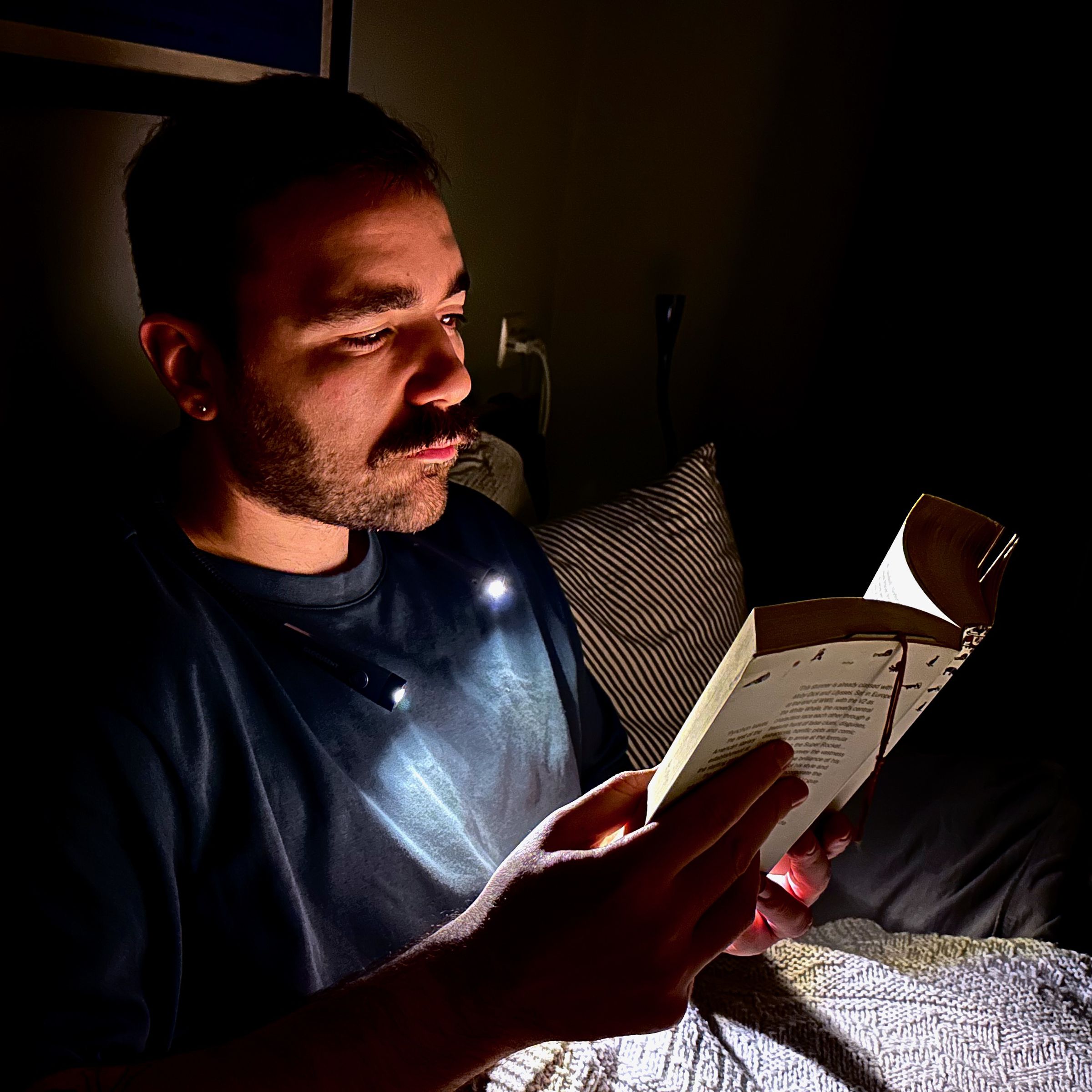 A man in a dark bedroom wears a neck lamp that illuminates just his face and the book he’s holding while sitting upright in bed. 
