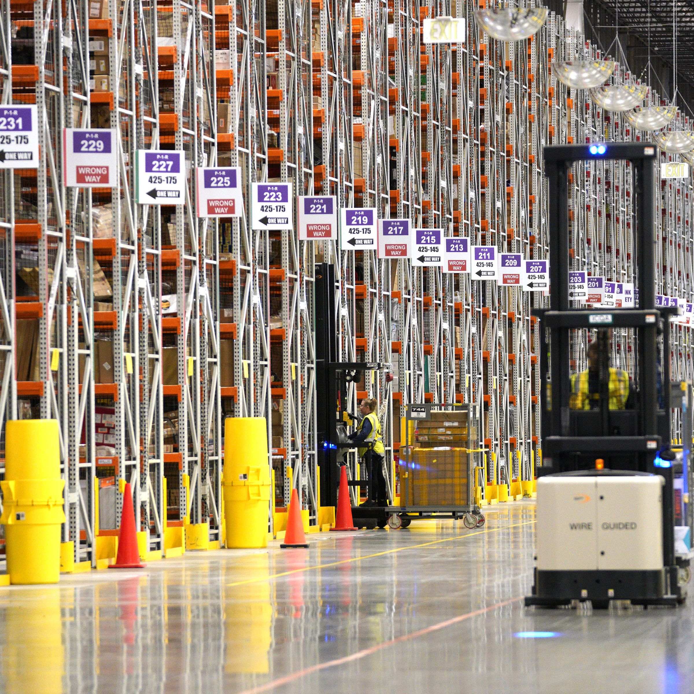 A worker drives a fork lift inside a warehouse past rows of shelves filled with packages.