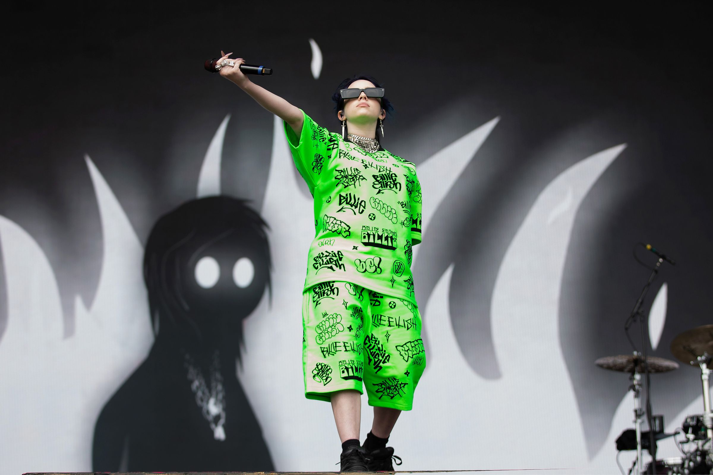 Billie Eilish stands onstage in front of a backdrop of greyscale flames and a white-eyed shillouette, holding a microphone outstretched towards the crowd