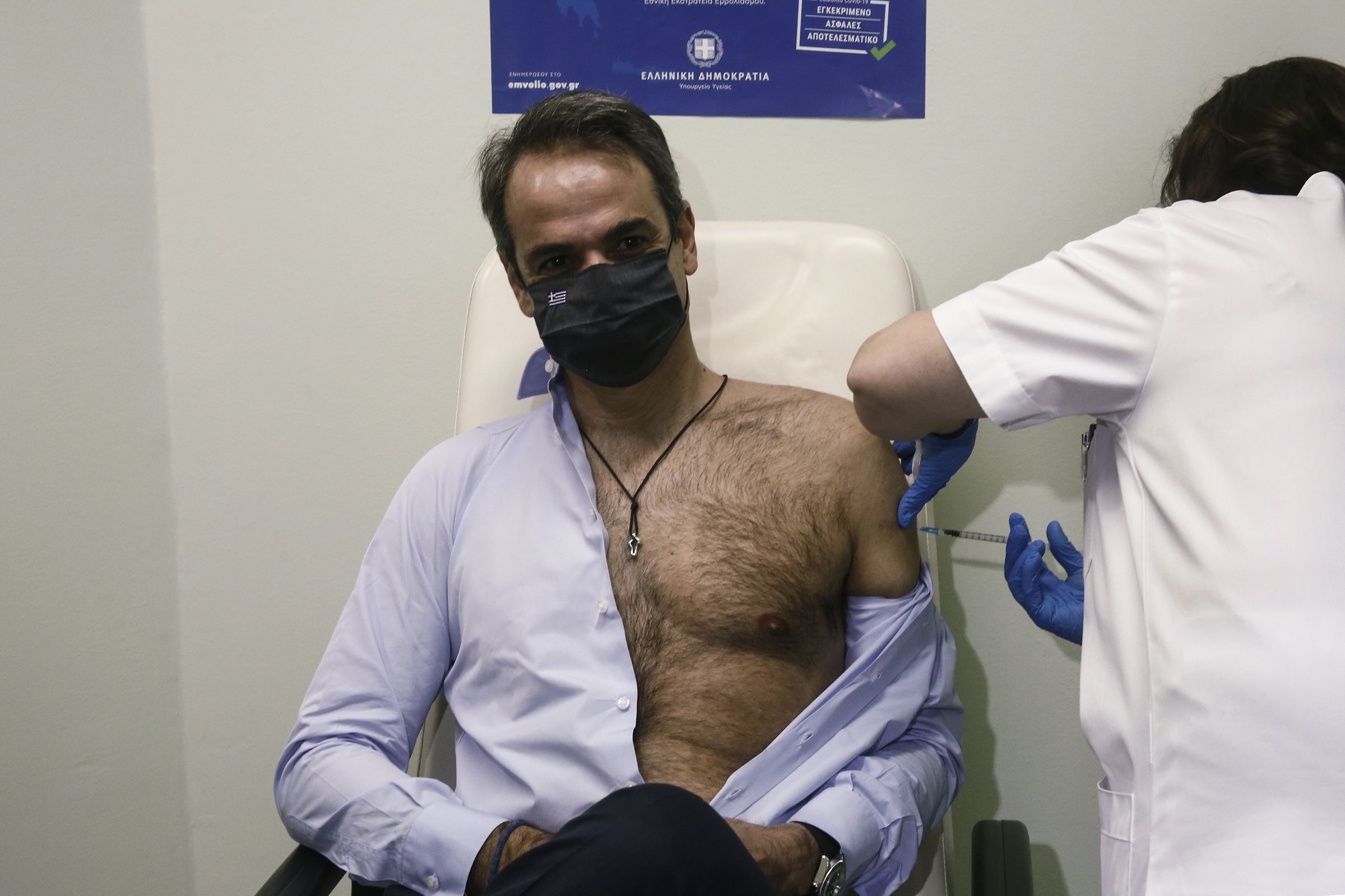 A man wearing a face mask sits in a chair with his shirt unbuttoned and half of his torso exposed as he gets the vaccine.