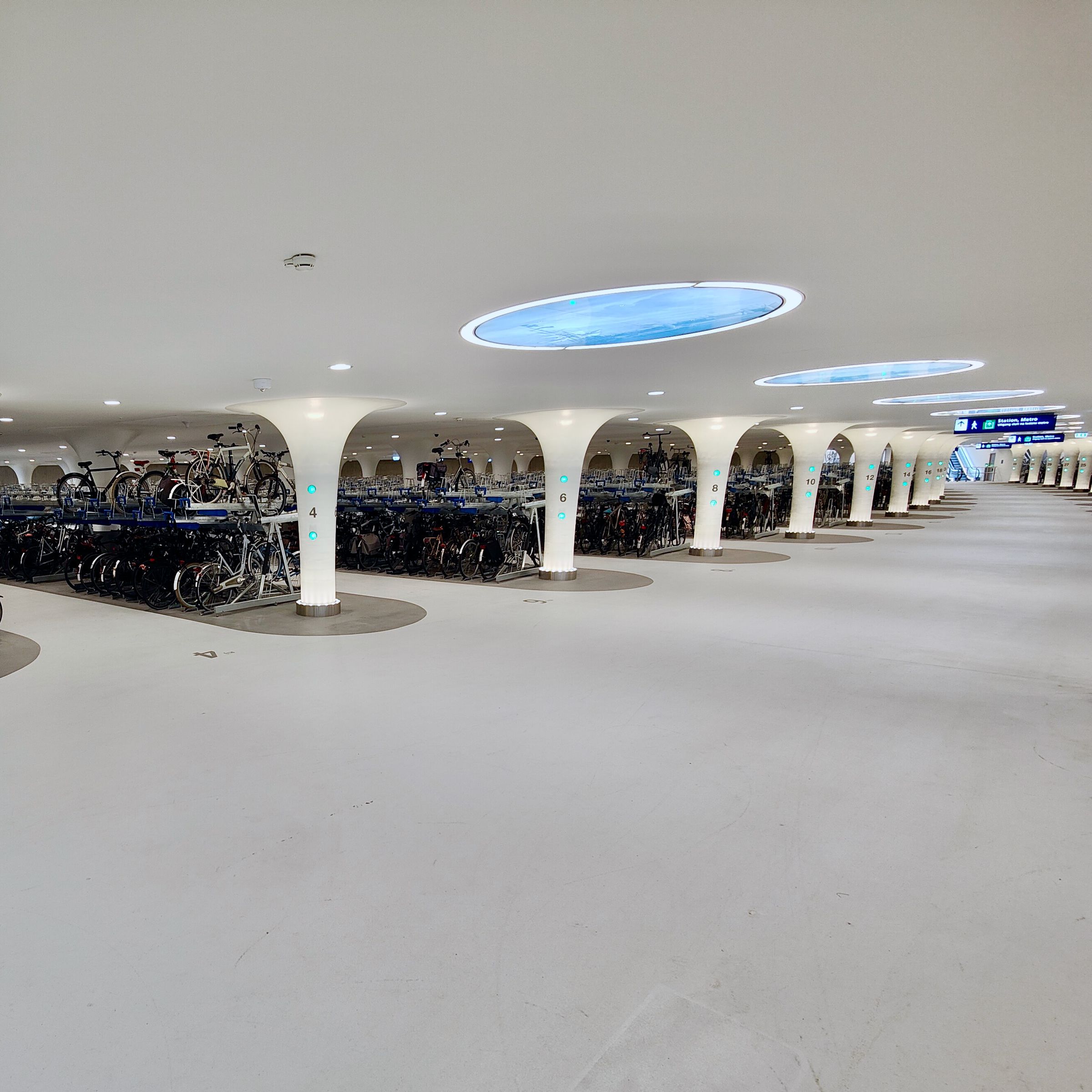 A photo showing a surprisingly spacious and futuristic parking garage of all white. Long rows of two-story bike racks extend to the edge of the room as tall white columns rise from the white floor to the white ceiling dotted with circular “windows” showing what looks like blue water (but are actually etched glass).