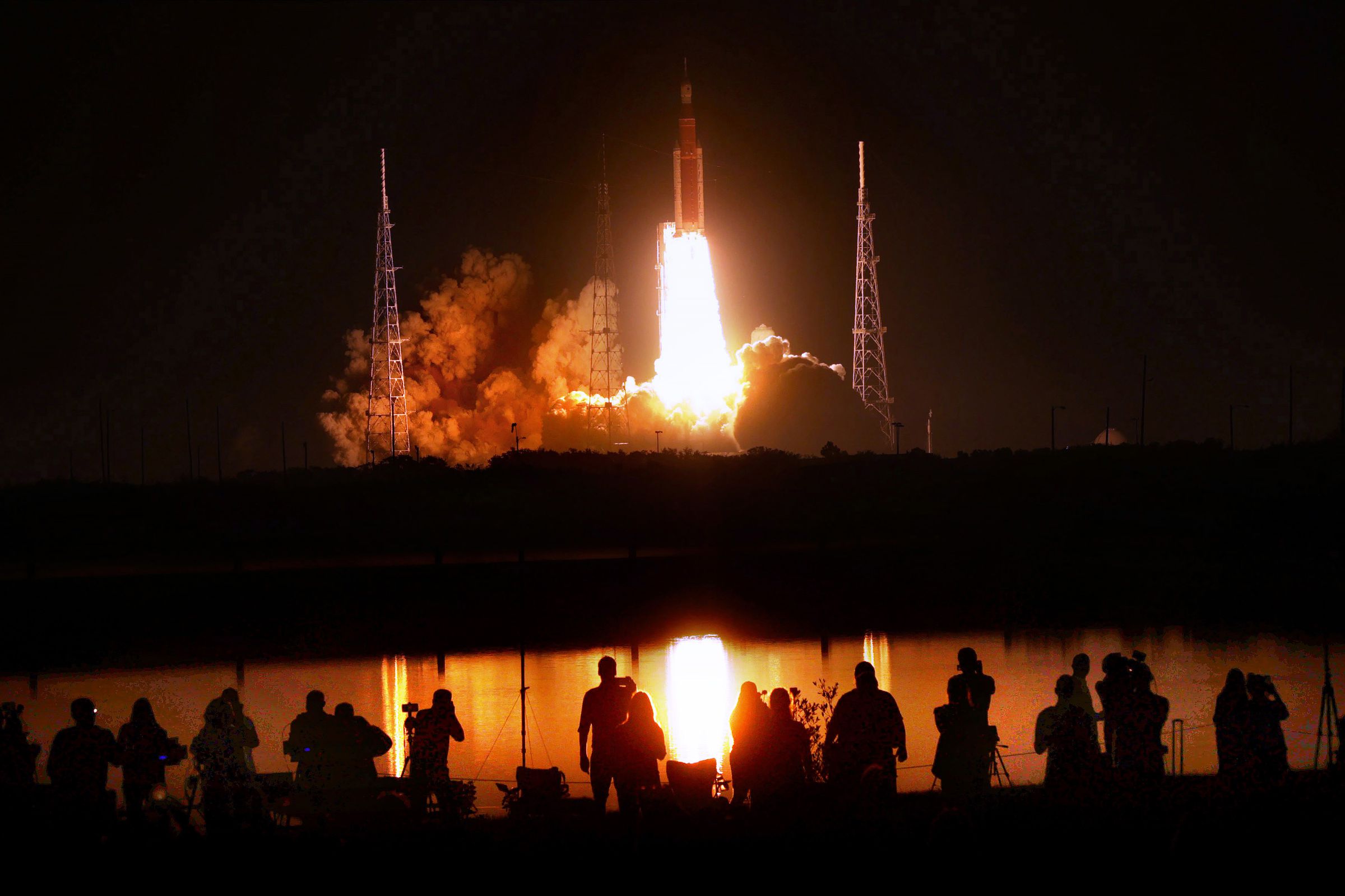 A row of people stand silhouetted against the orange glow of a distant rocket launch.
