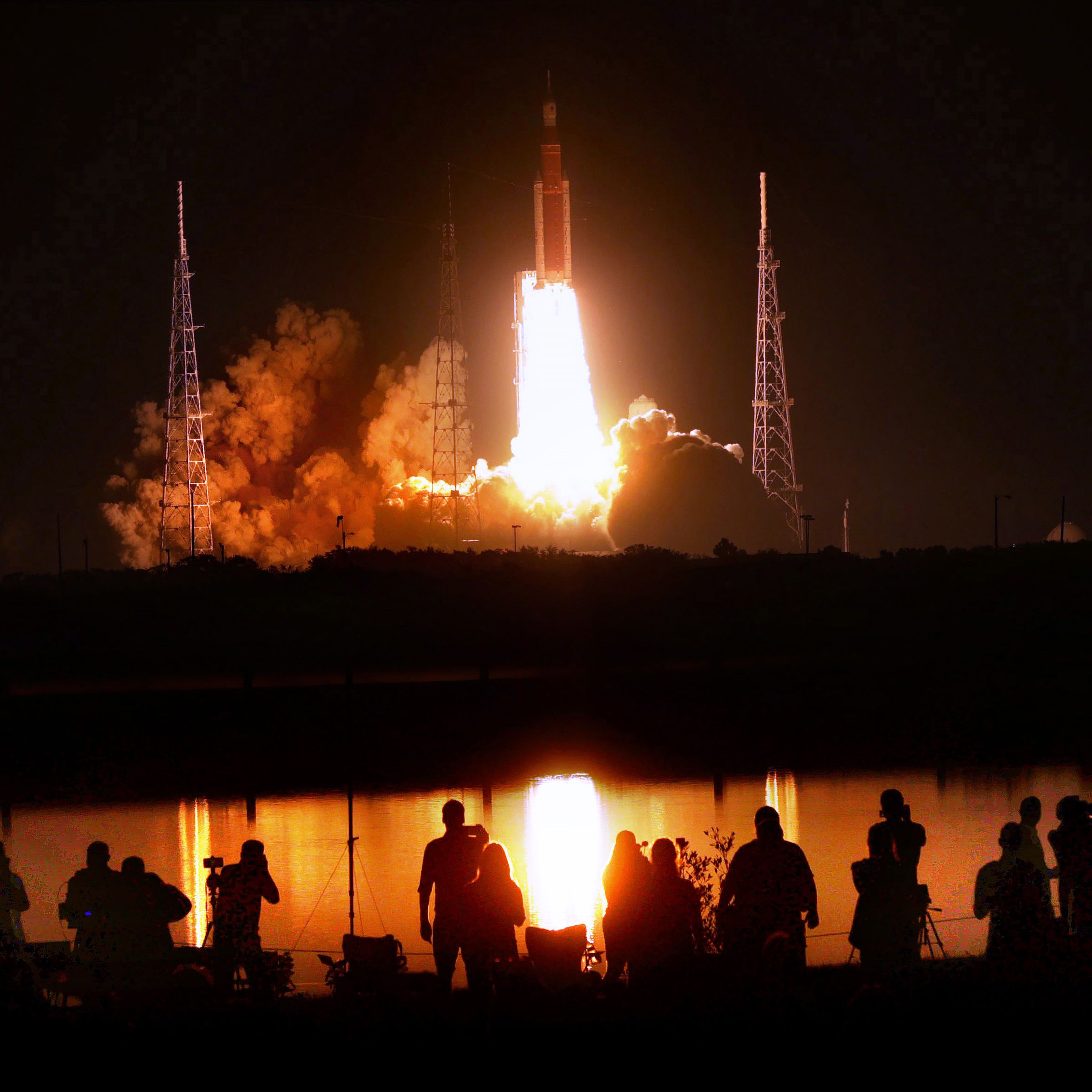 A row of people stand silhouetted against the orange glow of a distant rocket launch.