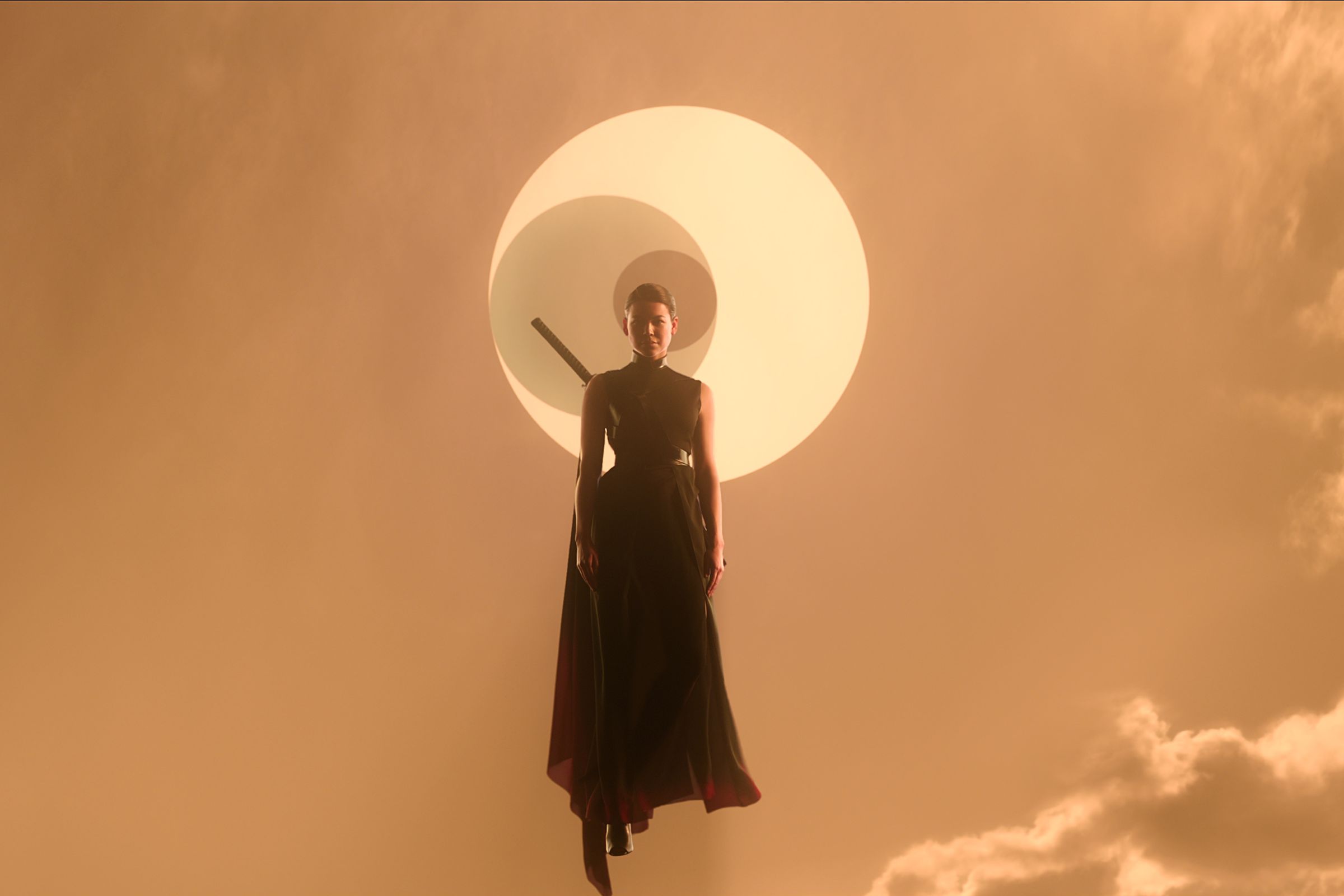 A woman in a form-fitting, sleeveless back outfit accented with a flowing black cape and a sheathed sword on her back. The woman is floating into a pale rust-colored sky in which the sun is being eclipsed by two smaller celestial bodies.