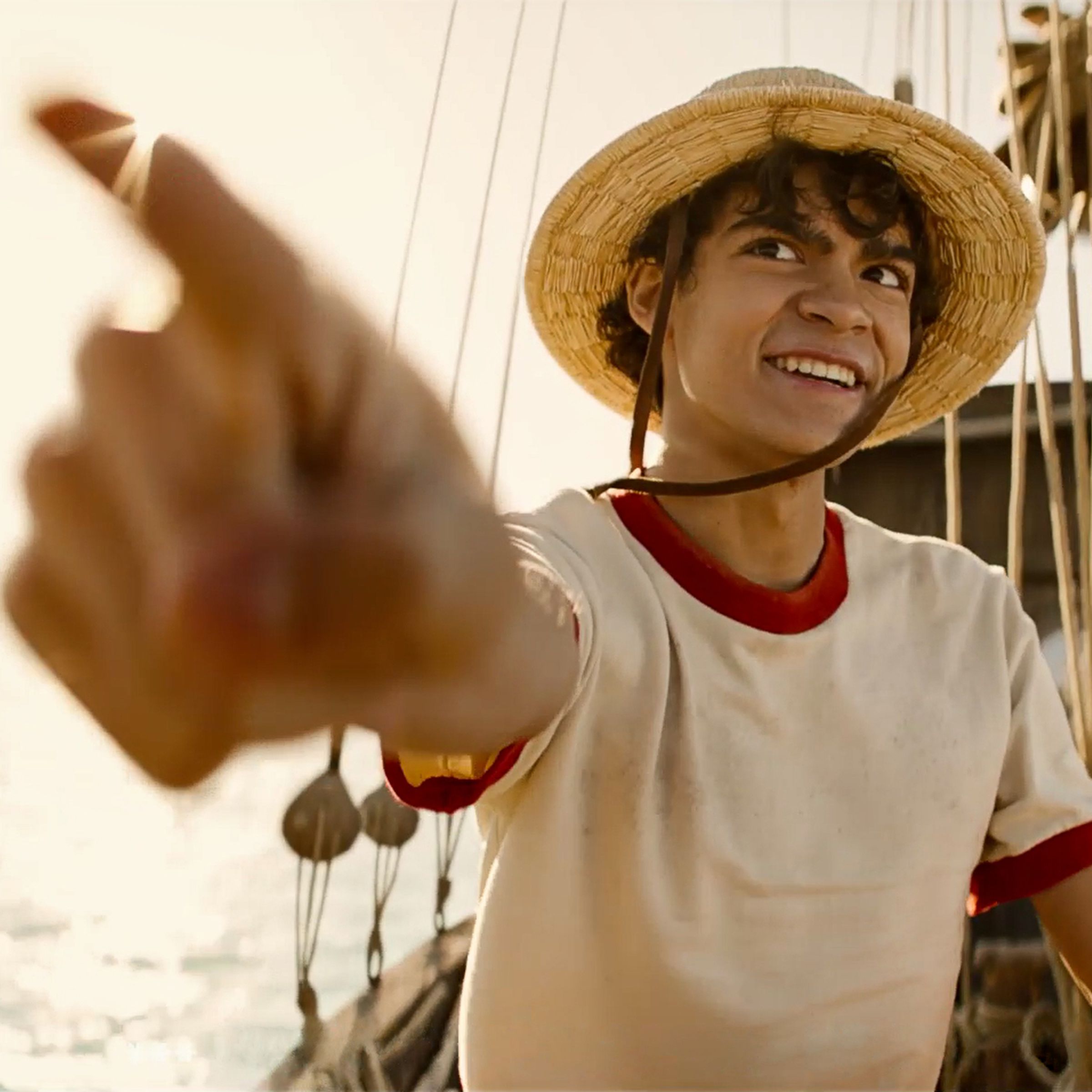 A boy wearing a straw hat and a white t-shirt ringed with red around the collar and sleeves. The boy is smiling, pointing towards the camera, and standing on the deck of a boat.