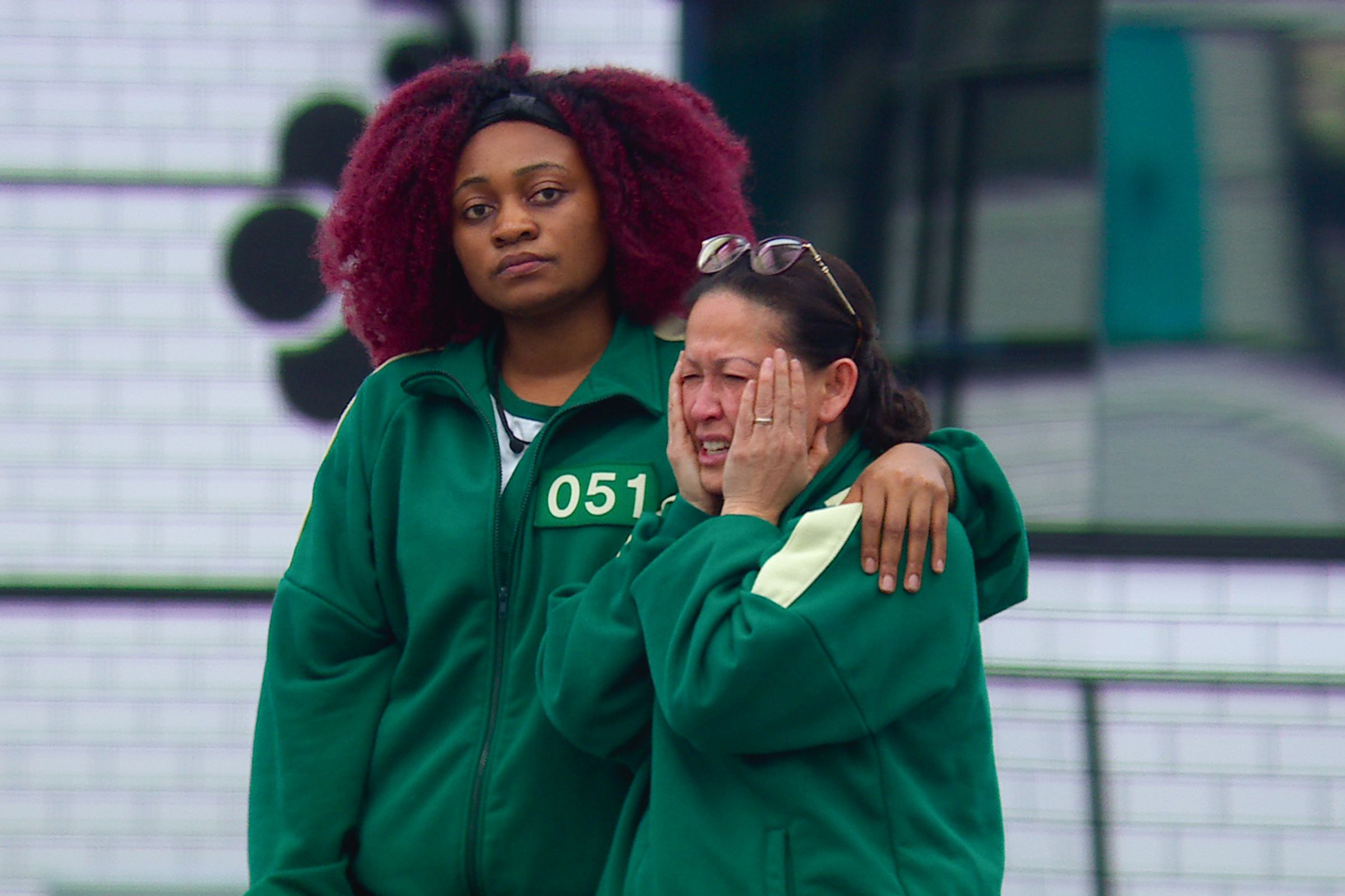 Two contestants wearing green jumpsuits stand together, with one appearing to be upset.