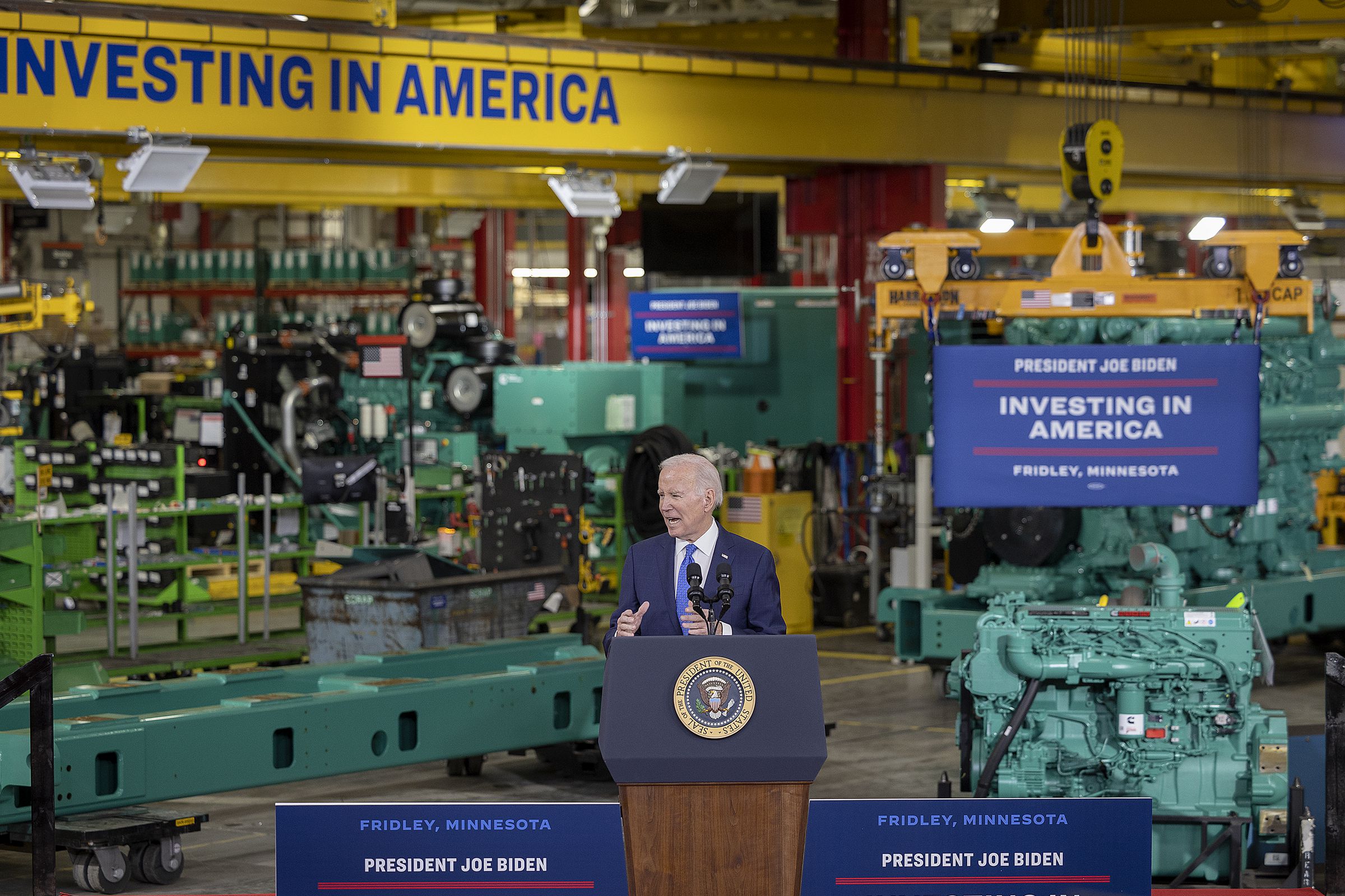 Joe Biden stands at a podium inside an industrial facility. Several signs around him say “Investing in America.”