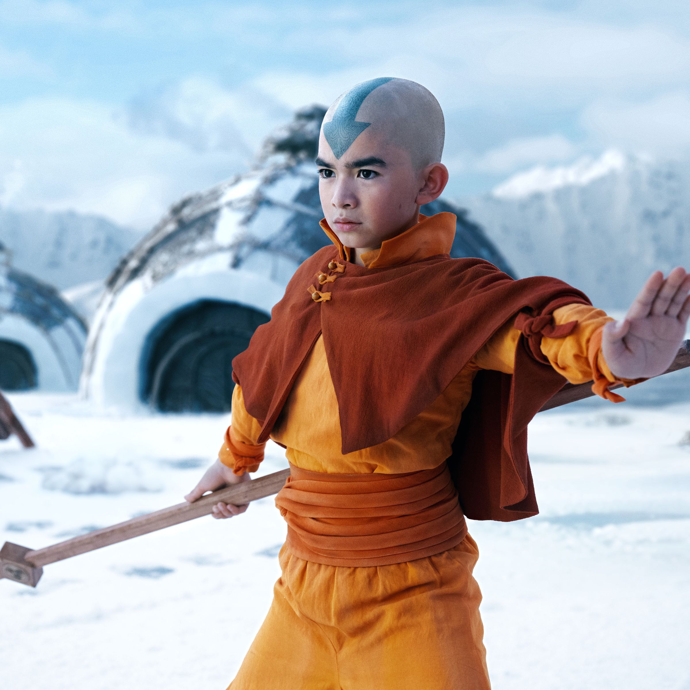 A bald boy whose head is adorned with a large, blue arrow tattoo. The boy is wearing an orange jumpsuit-like outfit accented with a short cape. The boy is also holding a wooden staff, holding out his free hand in defense, and standing in a frozen tundra.