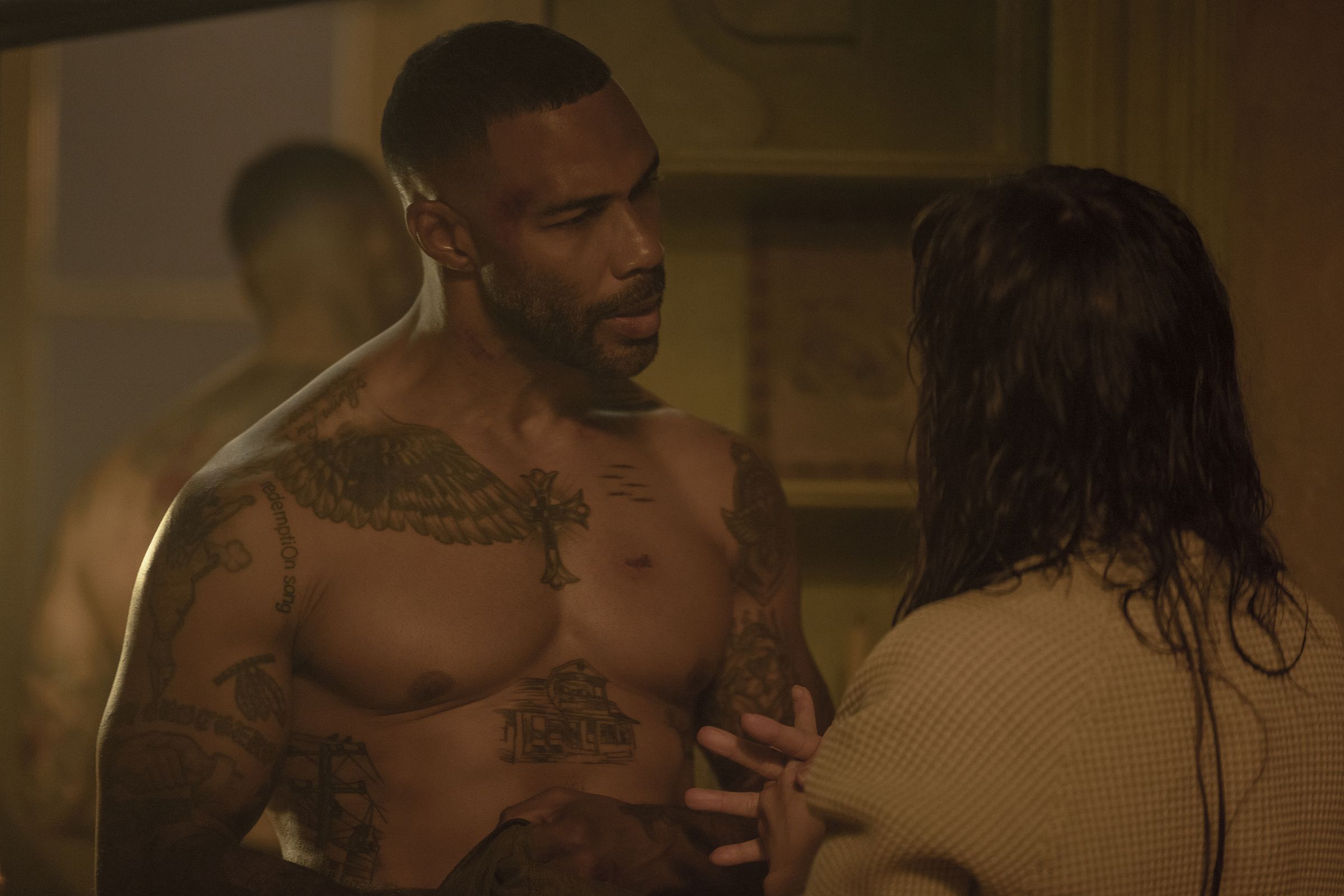 Omari Hardwicke, a Black Man, is wearing no shirt, showing off his physique and tattoos. Jennifer Lopez is speaking to him, but only the back of her head is visible.