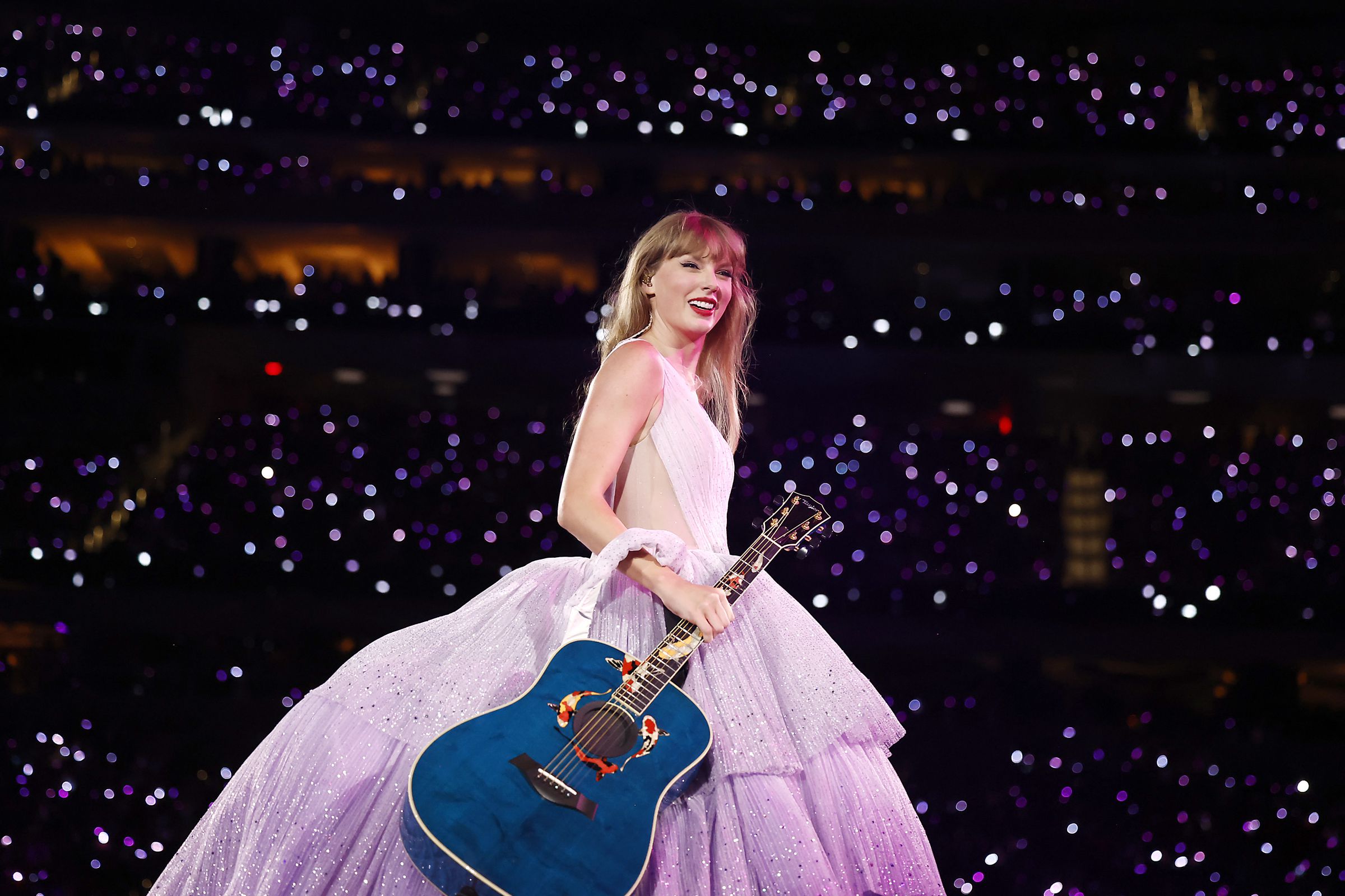 Taylor Swift holds a guitar on stage at the Eras Tour with lots of people behind her.