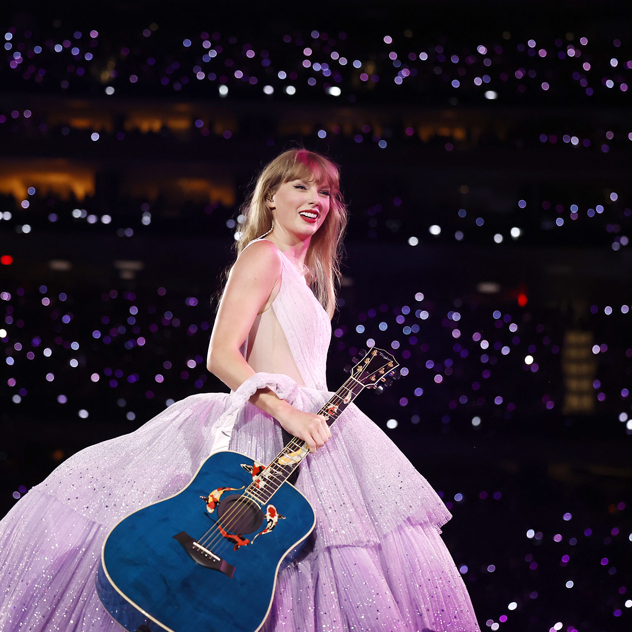 Taylor Swift holds a guitar on stage at the Eras Tour with lots of people behind her.