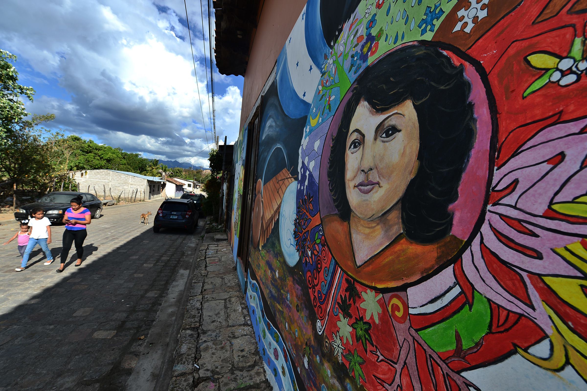 A woman and children walk next to a brightly painted wall with a mural depicting Berta Caceres.