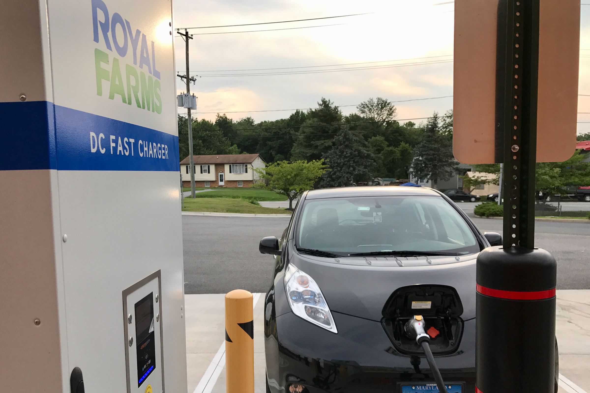 At sunset, a black Nissan Leaf is parked head on towards a Royal Farms DC Fast Charger. The CHAdeMO plug is already in the Leaf with its front latch open. There’s a small suburban house with many trees around in the background with an empty convenience store parking lot in the foreground.