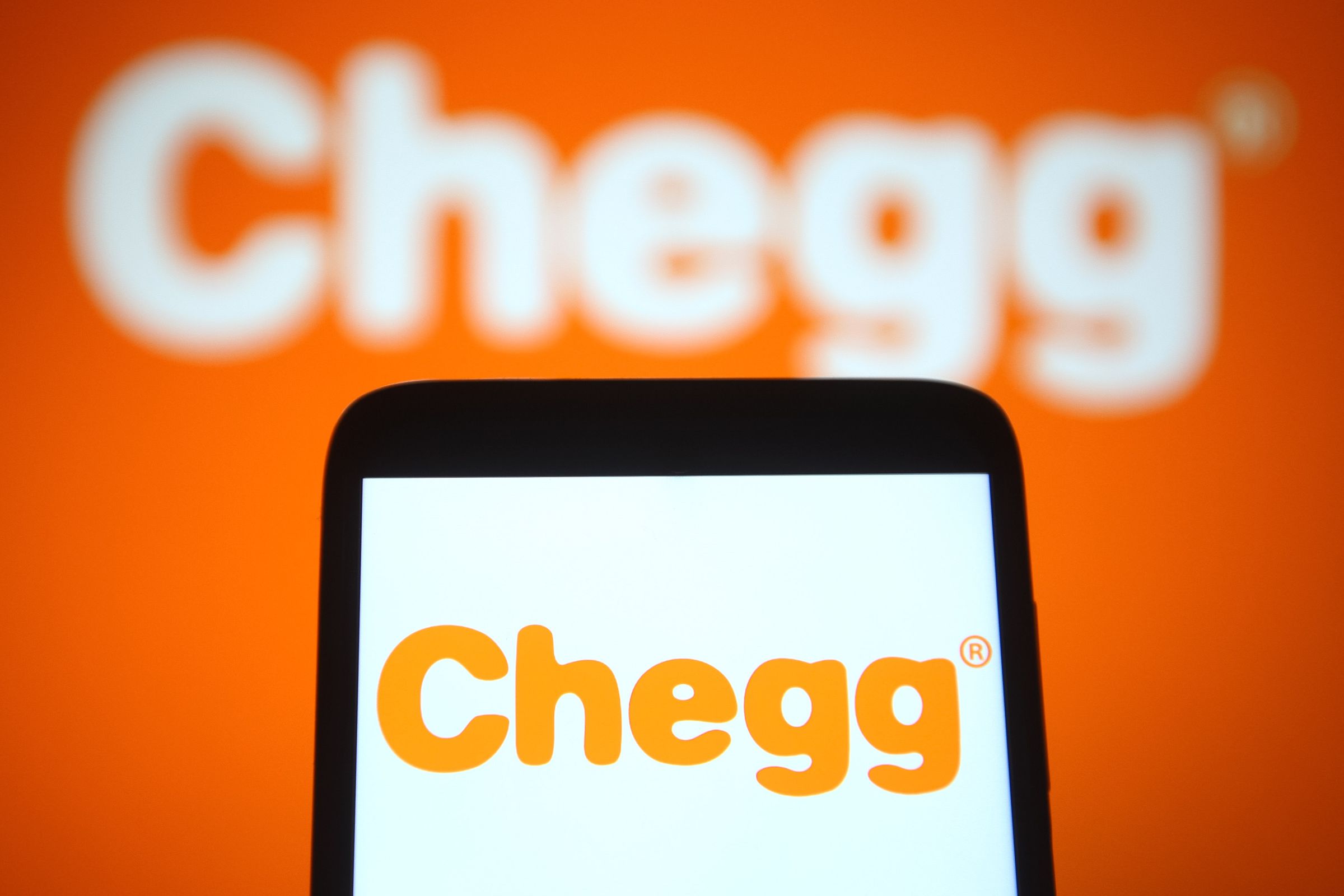 A Chegg, Inc. logo is displayed on both a mobile phone and on an orange wall behind the phone. 