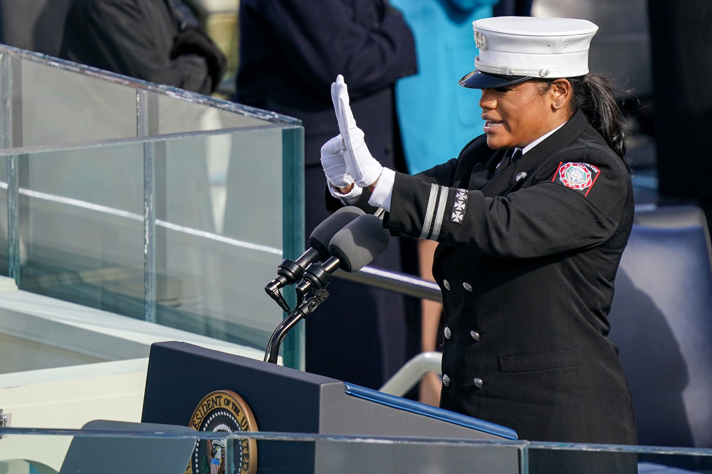 A Black woman in uniform stands at a podium, holding up her hands in the middle of signing.
