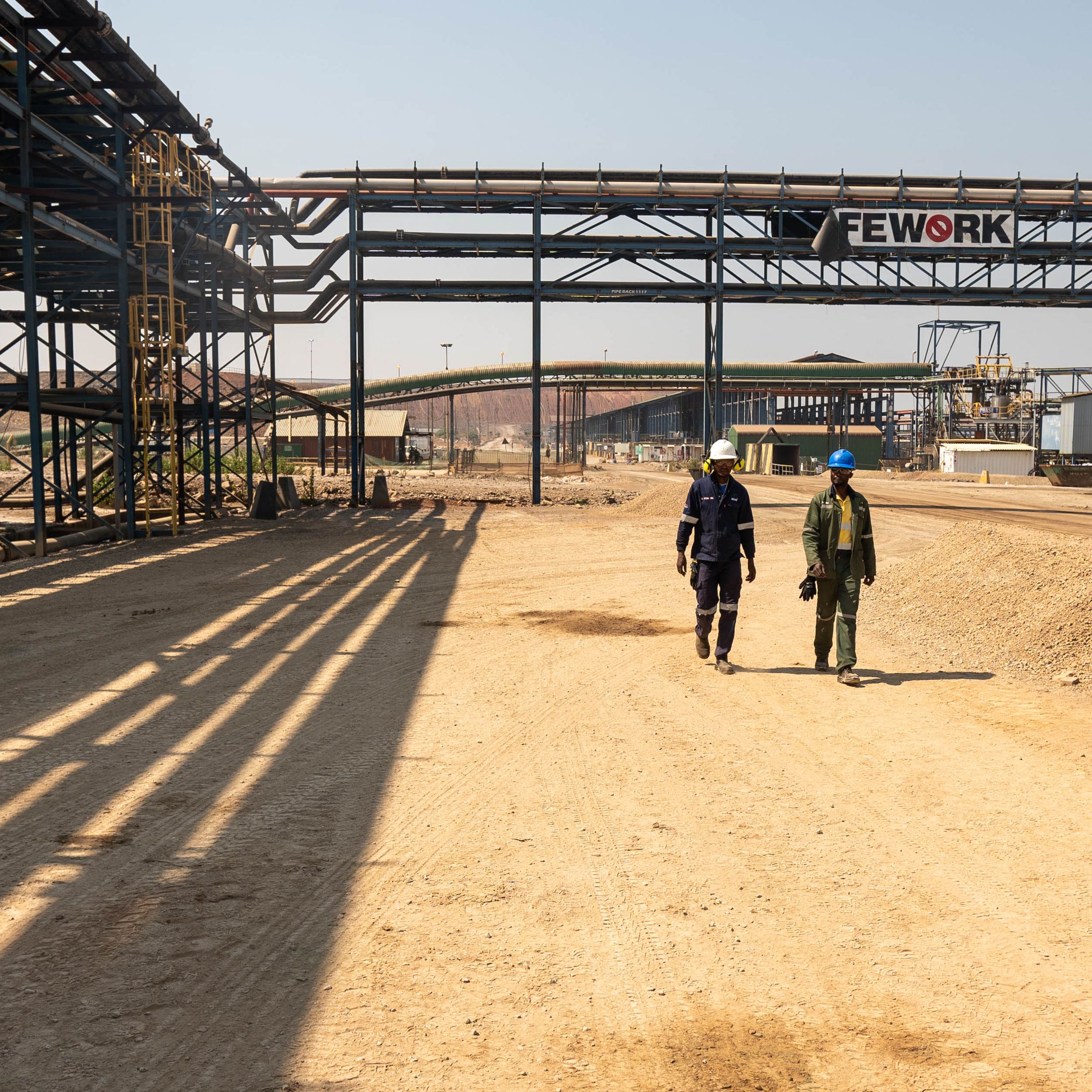 Two workers wearing hard hats walk through an industrial area.
