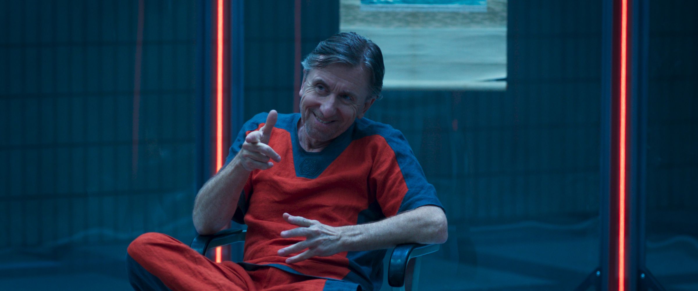 Tim Roth sitting in a plexiglass enclosure, sitting in a chair, and wearing a stylish prisoner’s uniform.