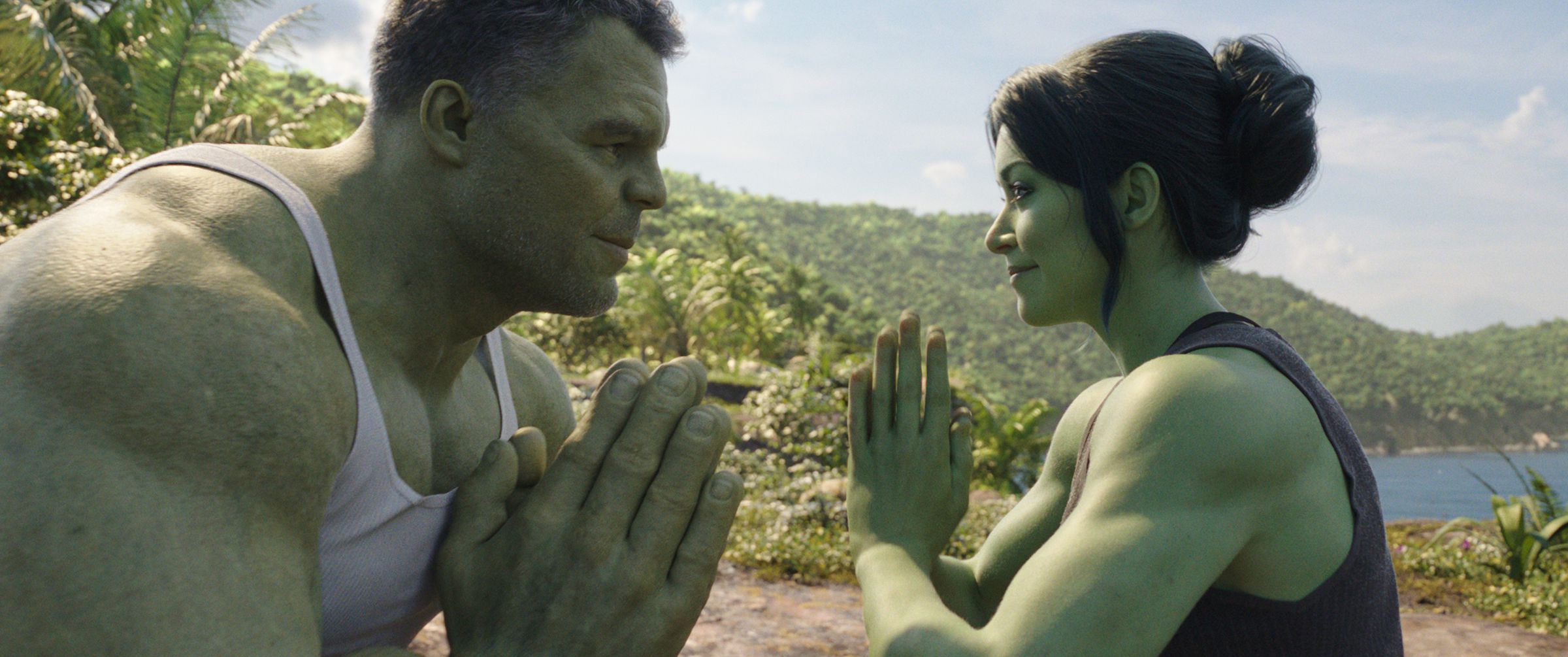 Smart Hulk wearing a white a-shirt sitting with his hands pressed together while he smiles at She-Hulk, who is sitting across from him in the same position, and wearing a black tank top. In the background, a remote island is pictured.