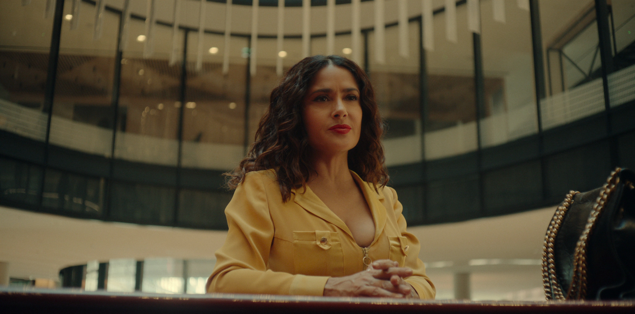 A Latina woman stands at a counter in a yellow jumpsuit. She looks annoyed.