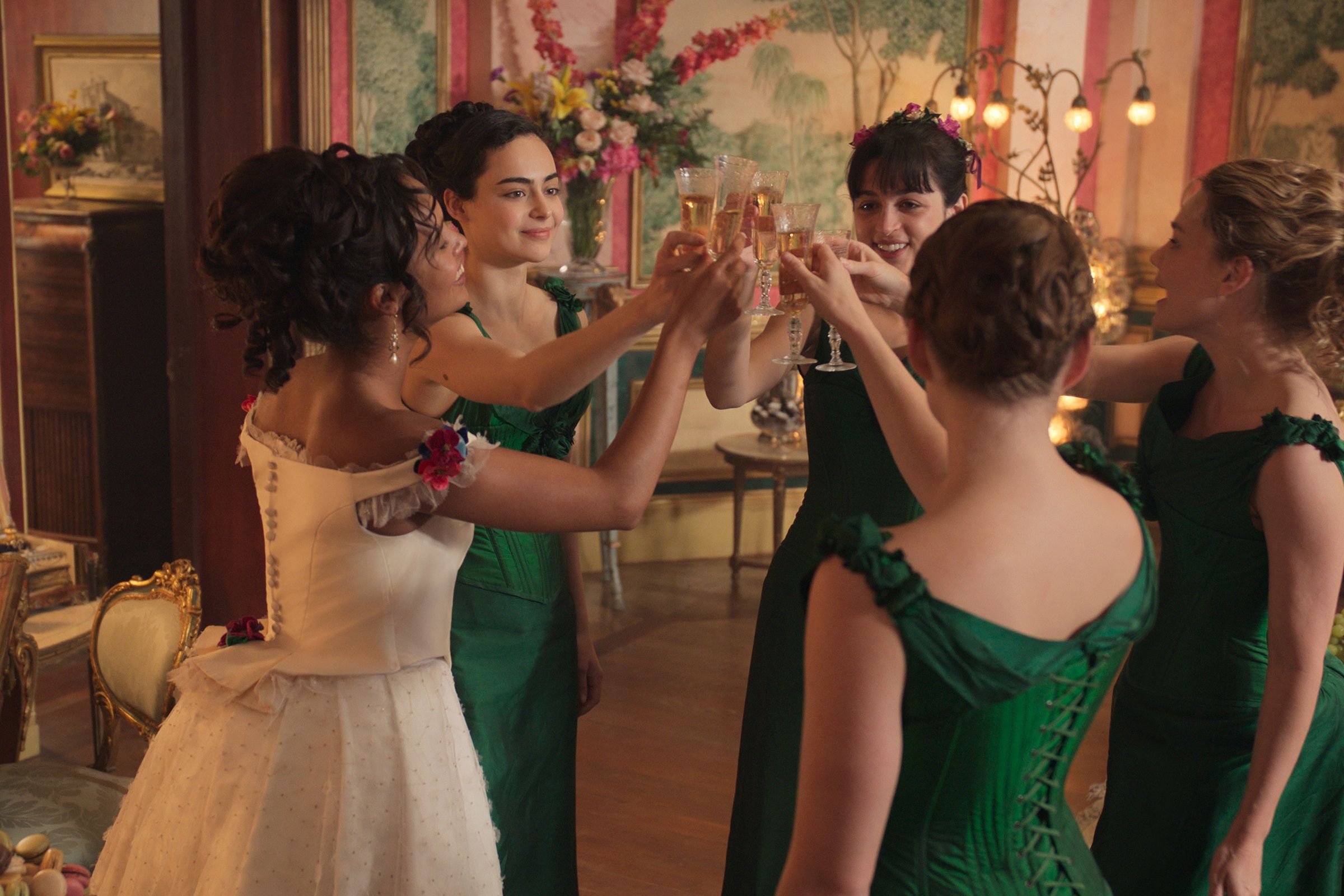 Five women, one in a white wedding dress and four in green bridesmaid dresses, toast in a very colorful and vibrantly pink room.