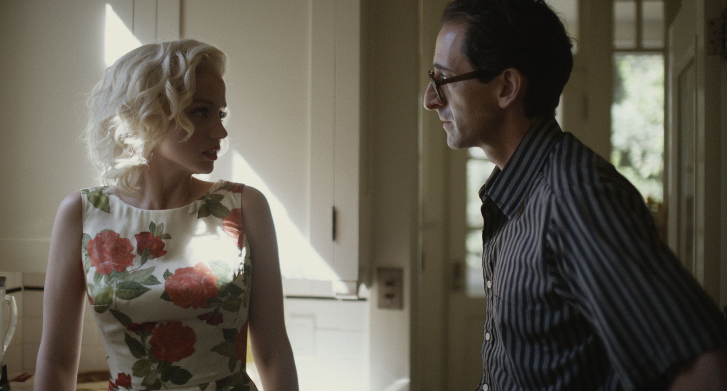 A blonde woman in a floral dress looking over in concern at a dark haired bespectacled man wearing a pinstripe shirt. The pair are standing in a well-lit but sparse kitchen.