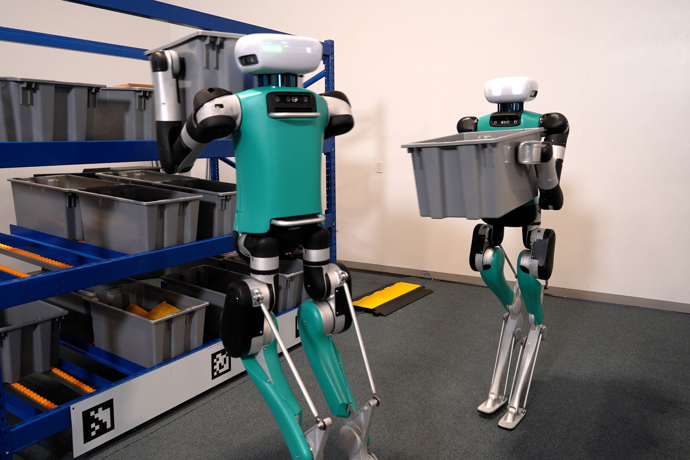 An image showing two robots in a warehouse, with one carrying a plastic bin and the other taking one off of a shelf
