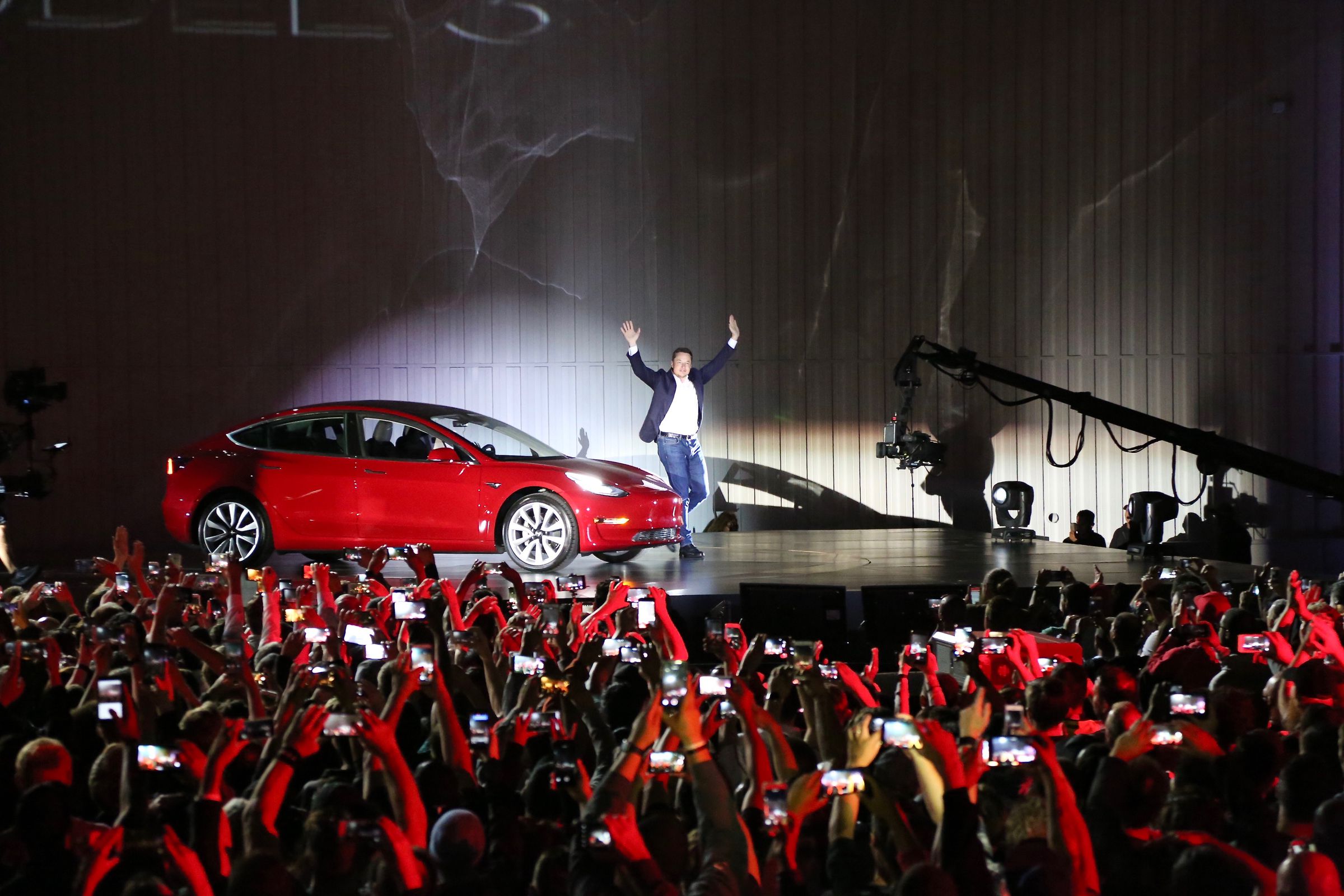 Elon Musk stands with his hands raised next to a red car on a stage in front of a crowd.
