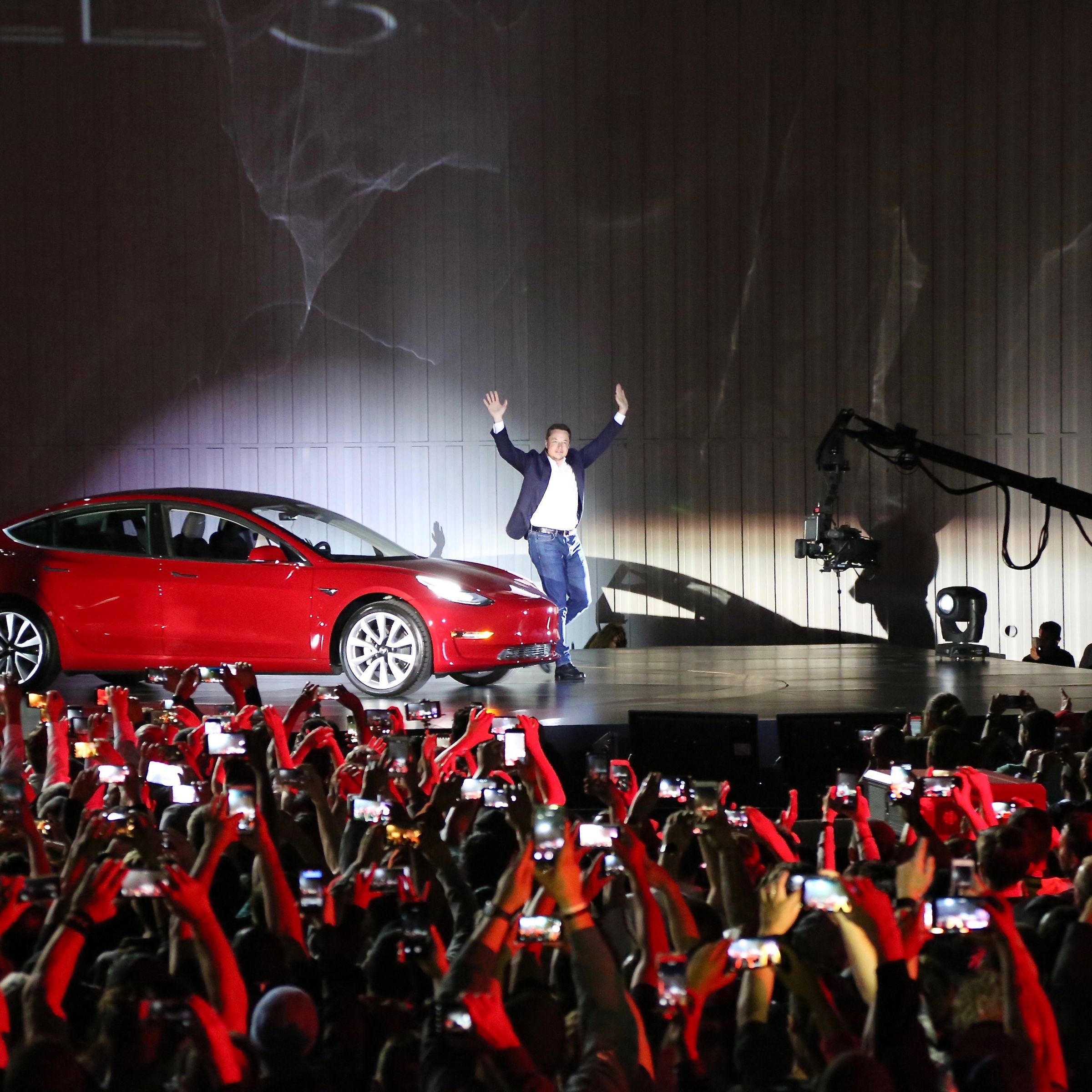 Elon Musk stands with his hands raised next to a red car on a stage in front of a crowd.