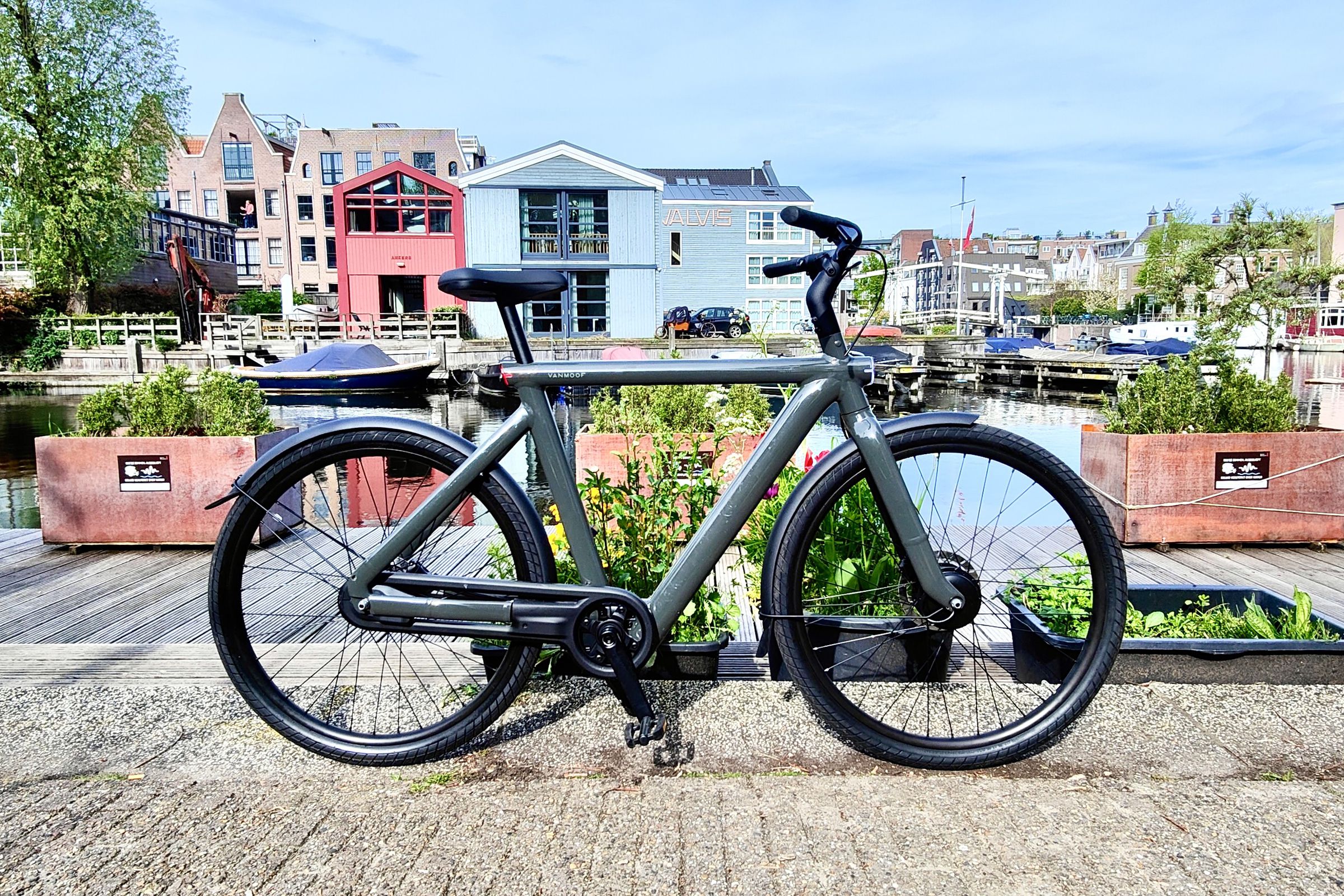 A dark gray electric VanMoof S5 bicycle stands in the foreground with modern Dutch homes, some light blue and red, in the background surrounded by water under a clear sky.