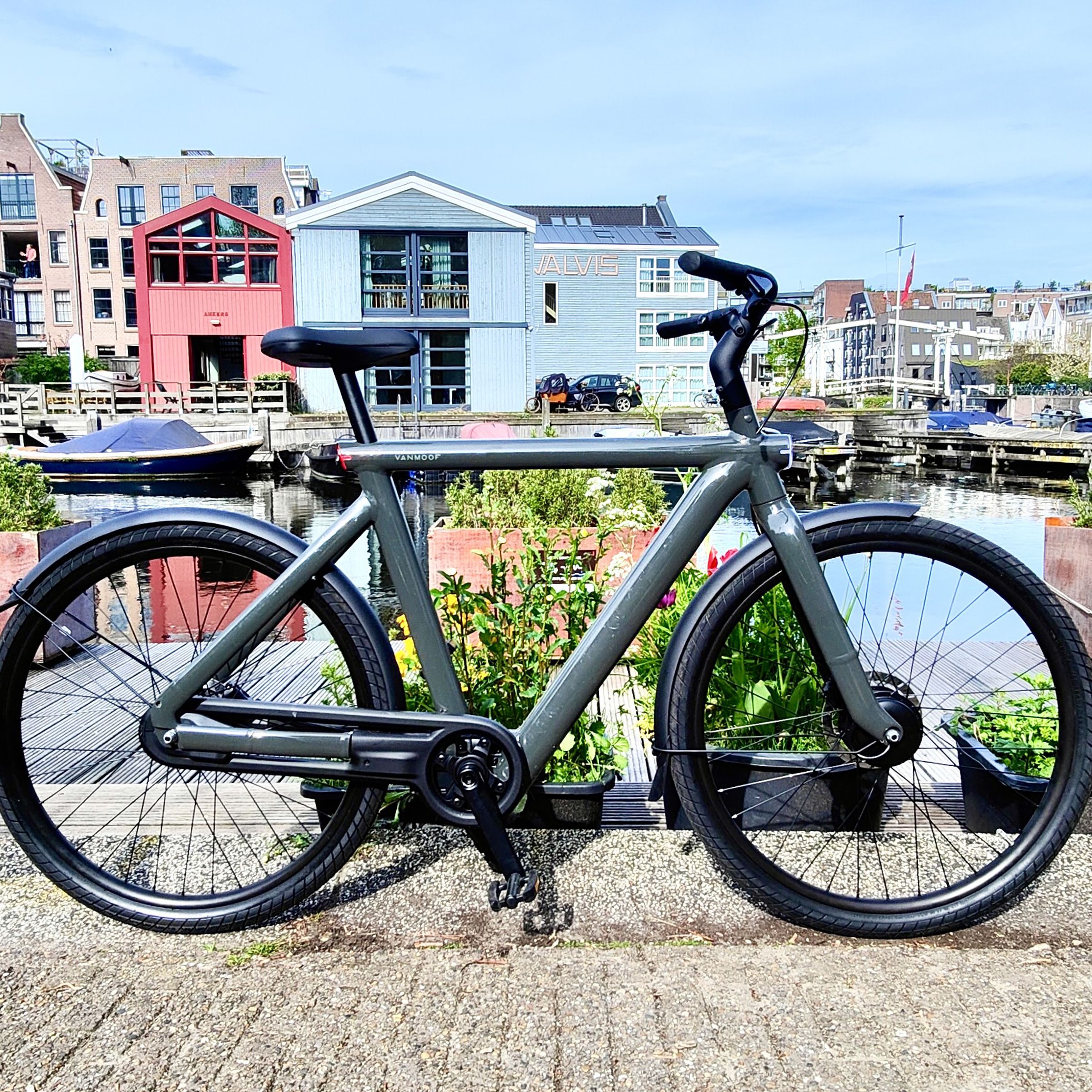 A dark gray electric VanMoof S5 bicycle stands in the foreground with modern Dutch homes, some light blue and red, in the background surrounded by water under a clear sky.
