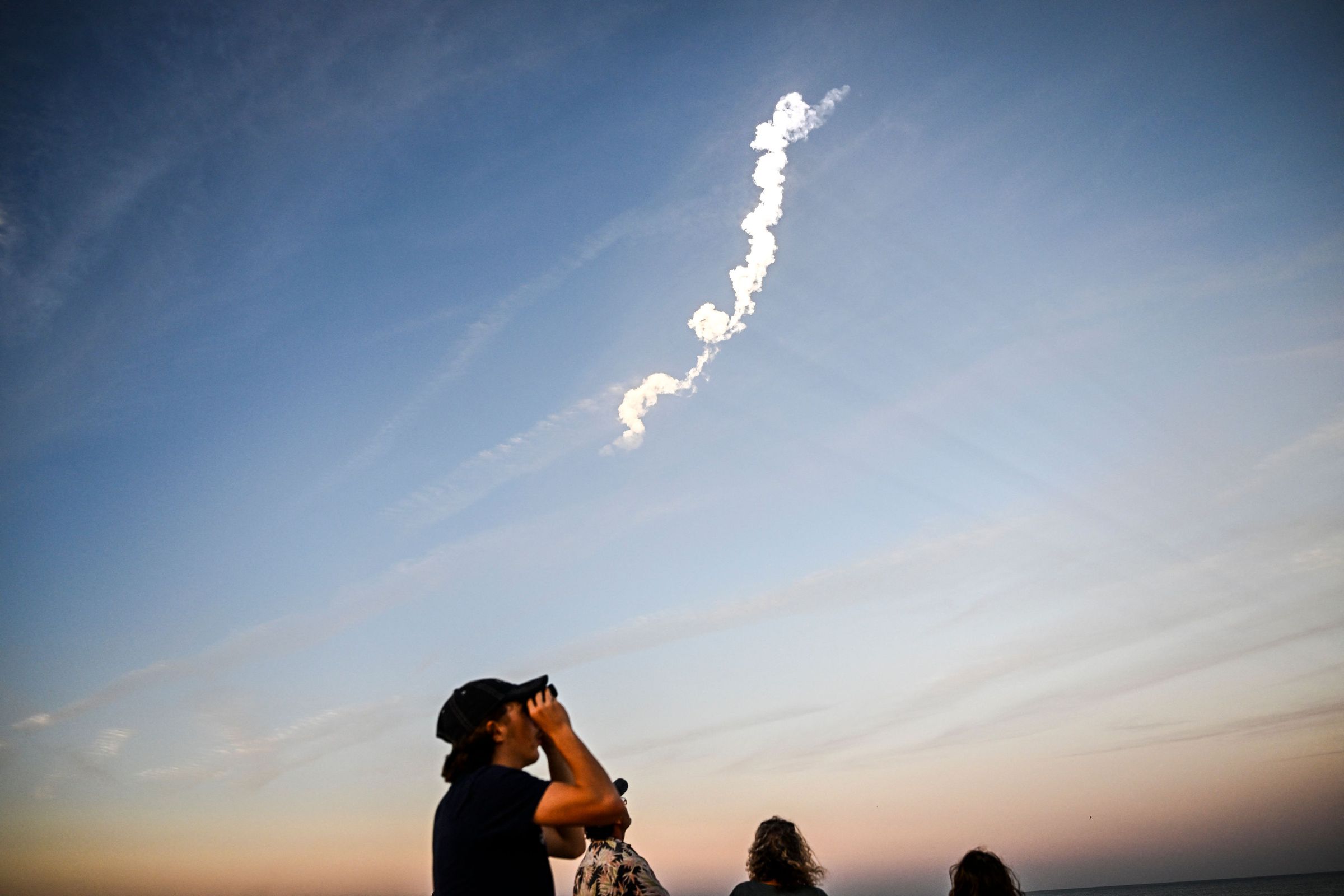 A person looks up into the sky at dusk with binoculars. There’s a rocket trail in the sky above the person.