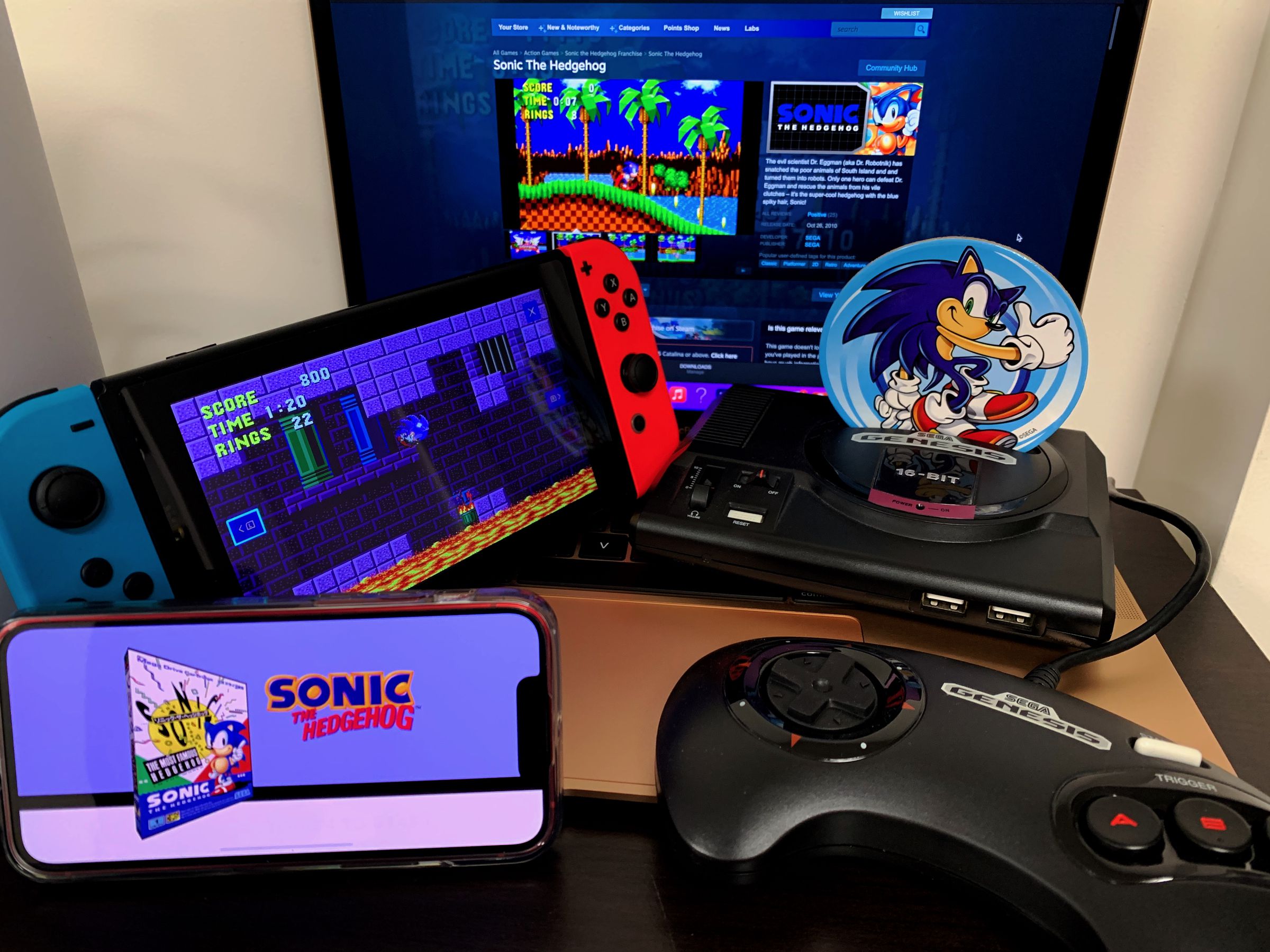 On a table, there are four devices: an iPhone 13 mini, Genesis mini, Nintendo Switch with blue and red joy cons, and a rose gold MacBook Air. devices with screens are showing sonic the hedgehog 1 game. A sonic character image on a coaster is propped up in the genesis console slot.