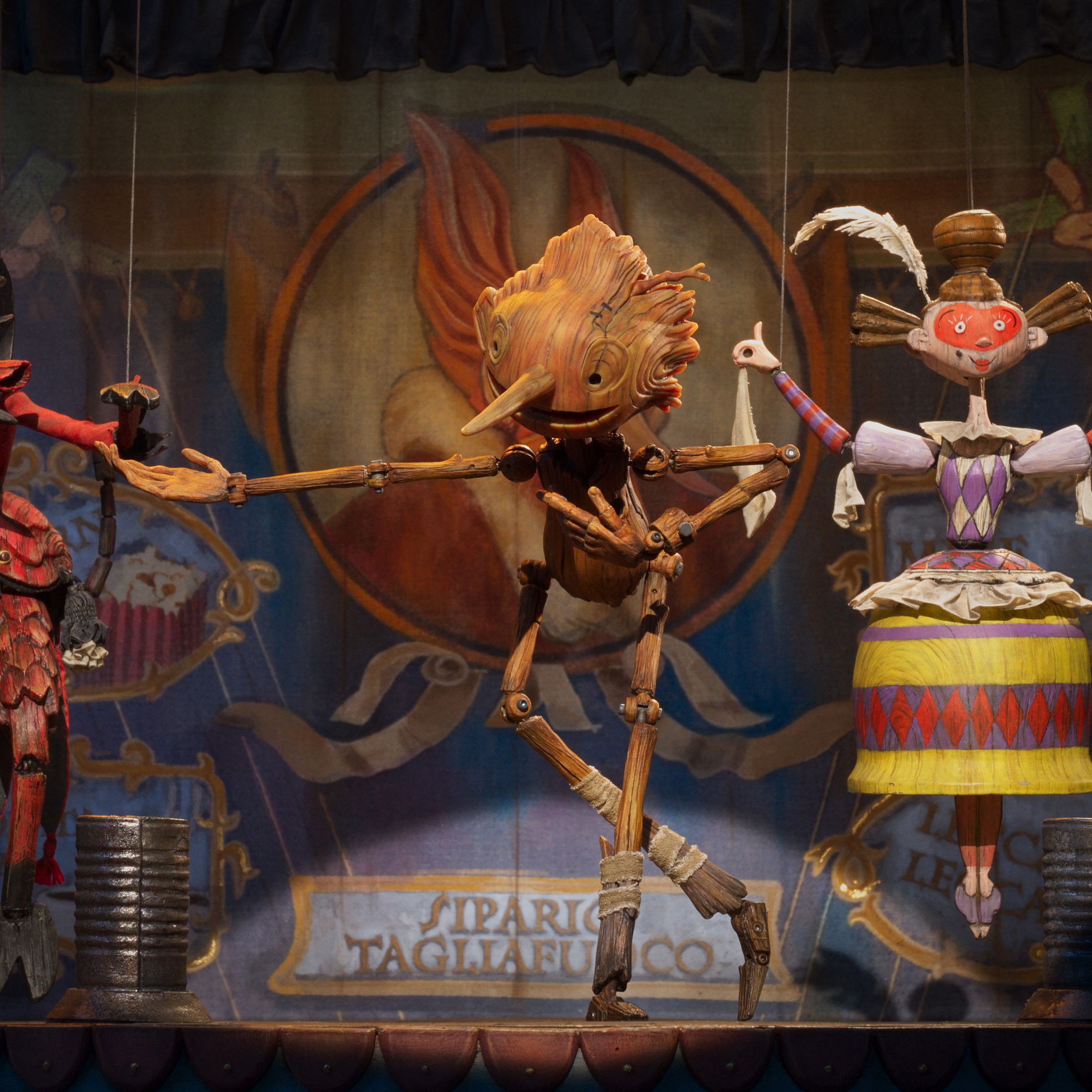 A wooden puppet shaped like a boy dancing on a small stage between a puppet made to resemble the devil and another resembling a dancer.