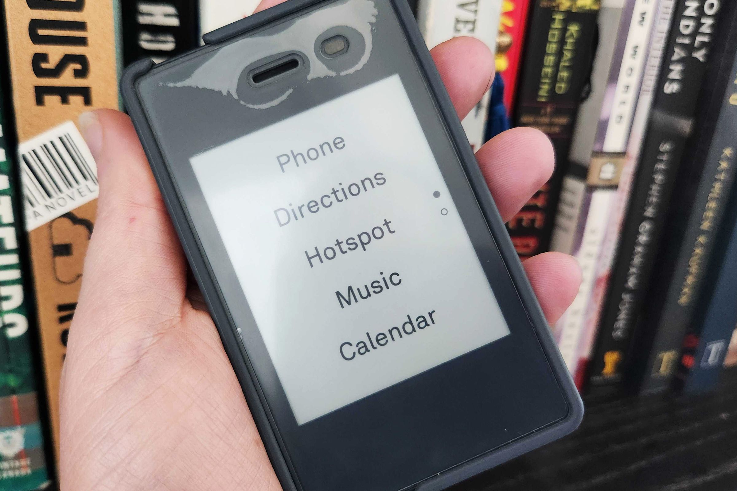 A small cellphone held in a person’s hand with books in the background.